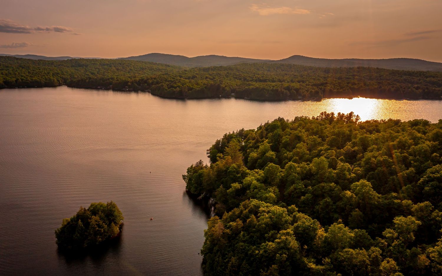 Aerial view of Cranberry Lake in the Adirondacks, New York, illuminated by golden sunset light with forested shoreline and calm water.