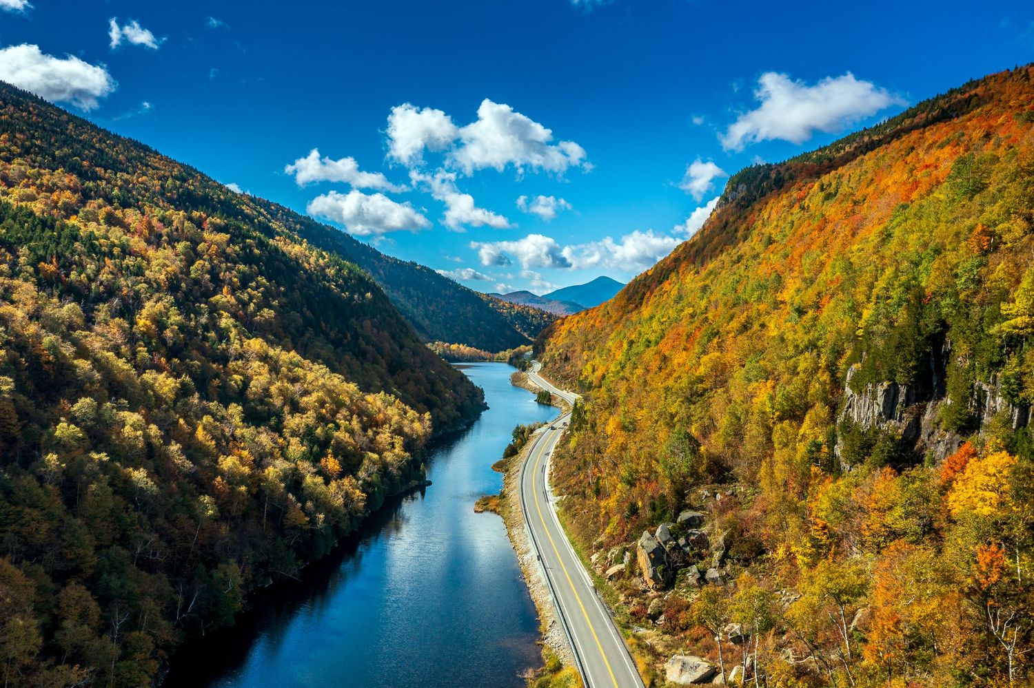 Aerial view of NY-73 winding along a reflective river between vibrant autumn mountains in the Adirondacks, New York, under a bright blue sky