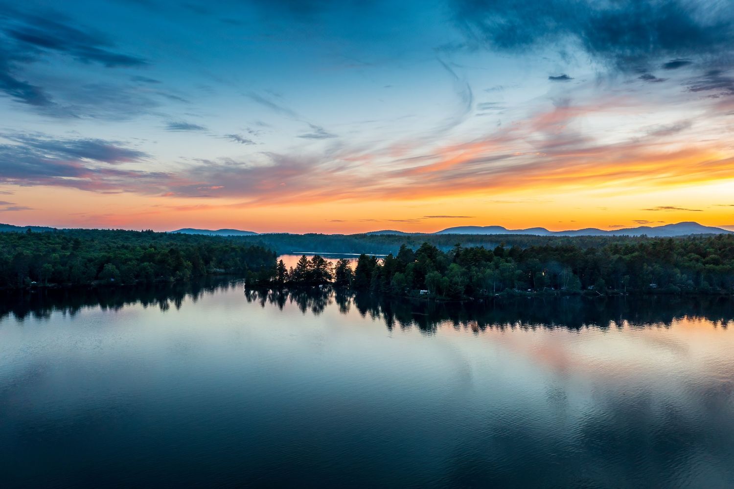 Vibrant sunset sky reflecting over a calm mountain lake in the Adirondacks, New York, surrounded by forested shores and distant peaks