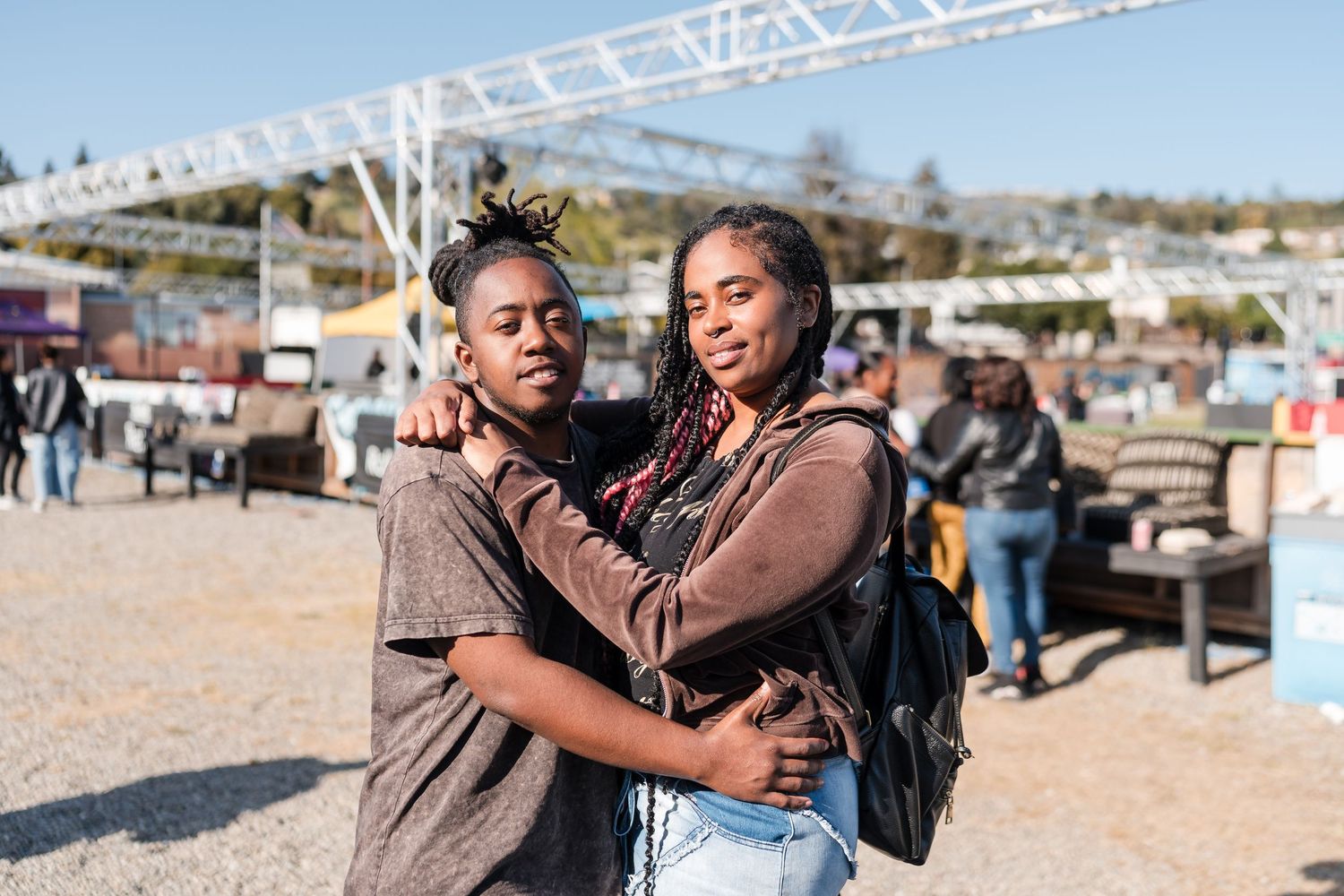 Two friends embrace warmly at an outdoor festival with event structures in the background.