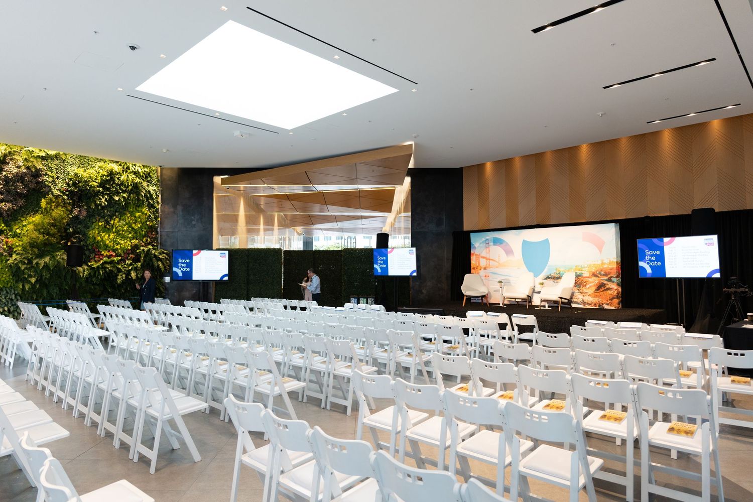 Modern conference room setup with rows of white folding chairs facing presentation screens and wooden stage area.