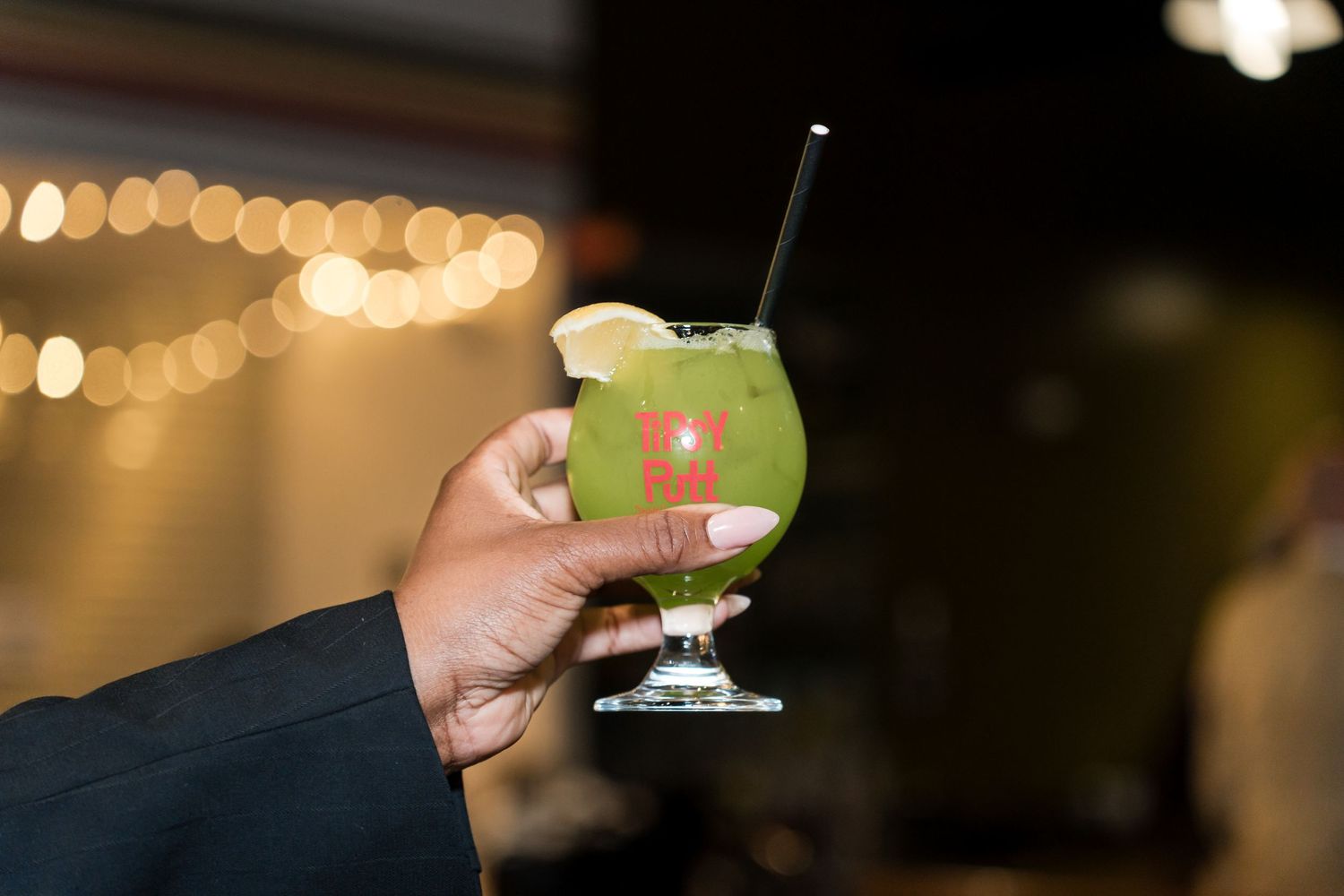 Hand holding a green cocktail in a martini glass with blurred bokeh lights in the background.