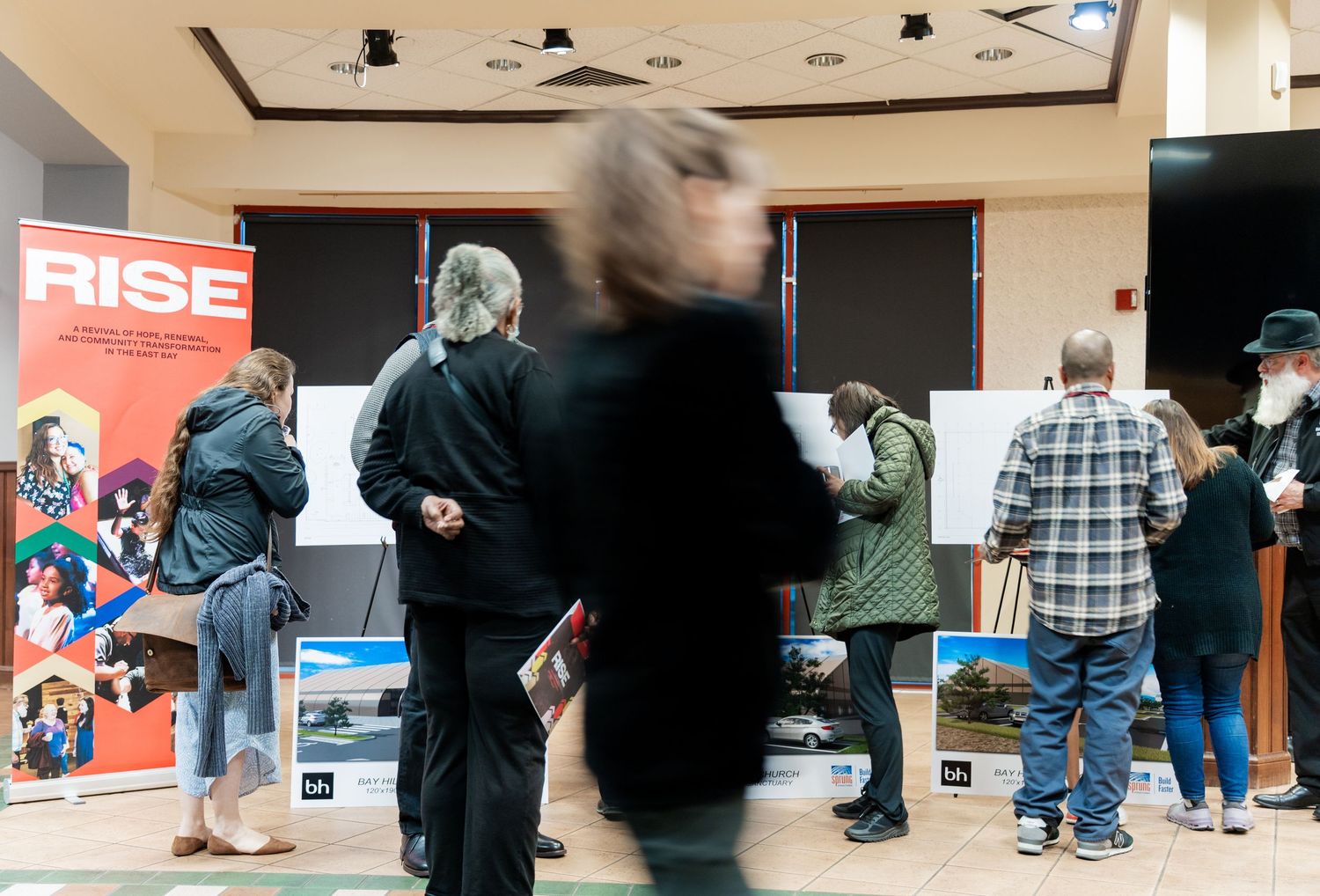 People gather in a line at an event venue with displays and signage visible in the background.