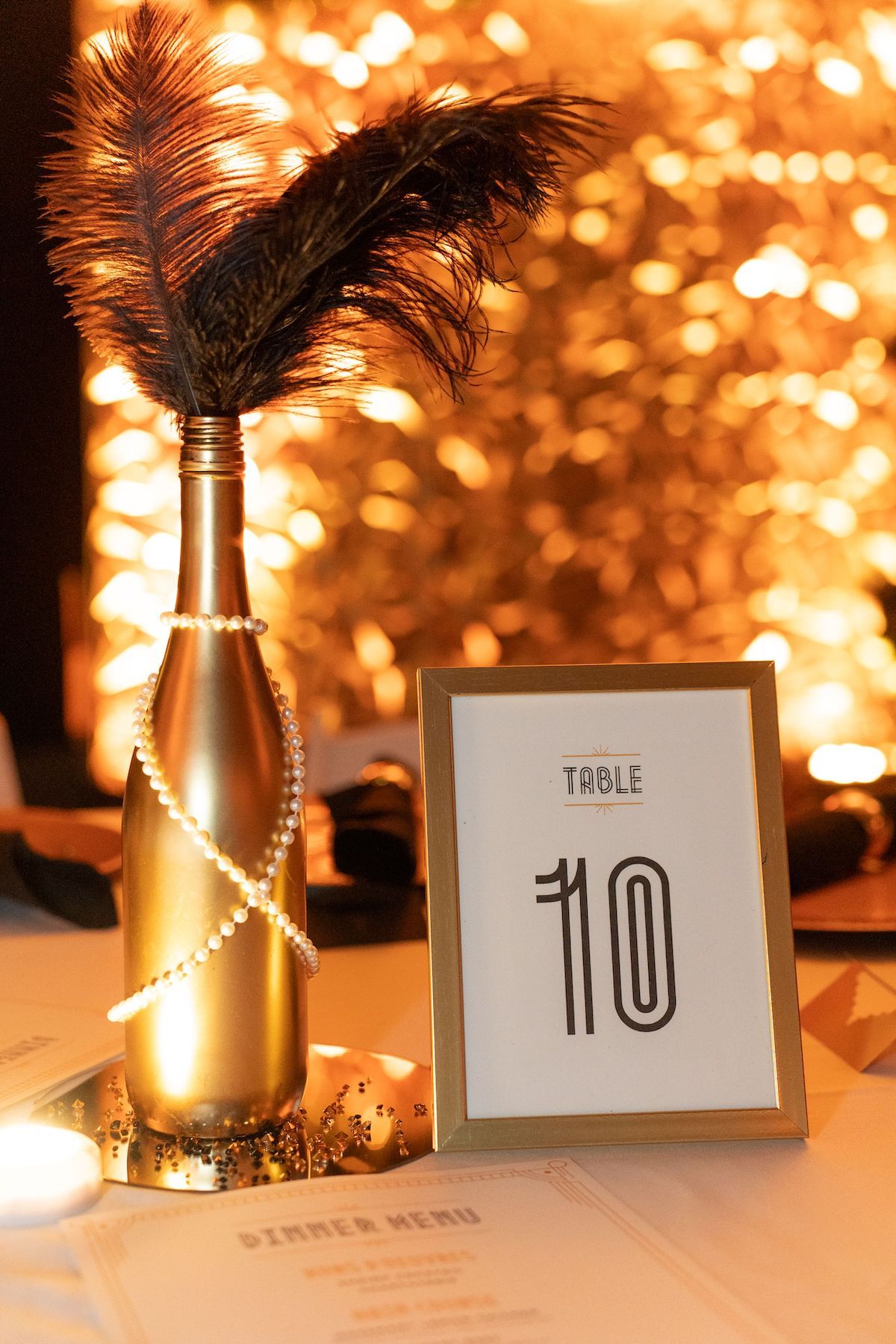 Elegant table number cards and silver vases with feathers displayed against a warm glowing bokeh background at an event.
