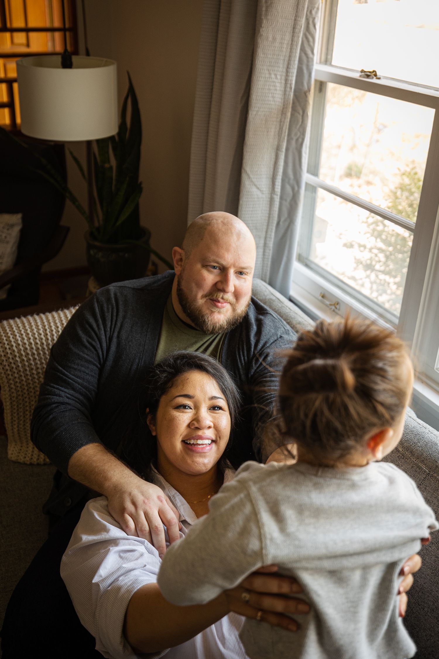 A smiling family of three sharing a warm moment on a couch near a window in a cozy living room.