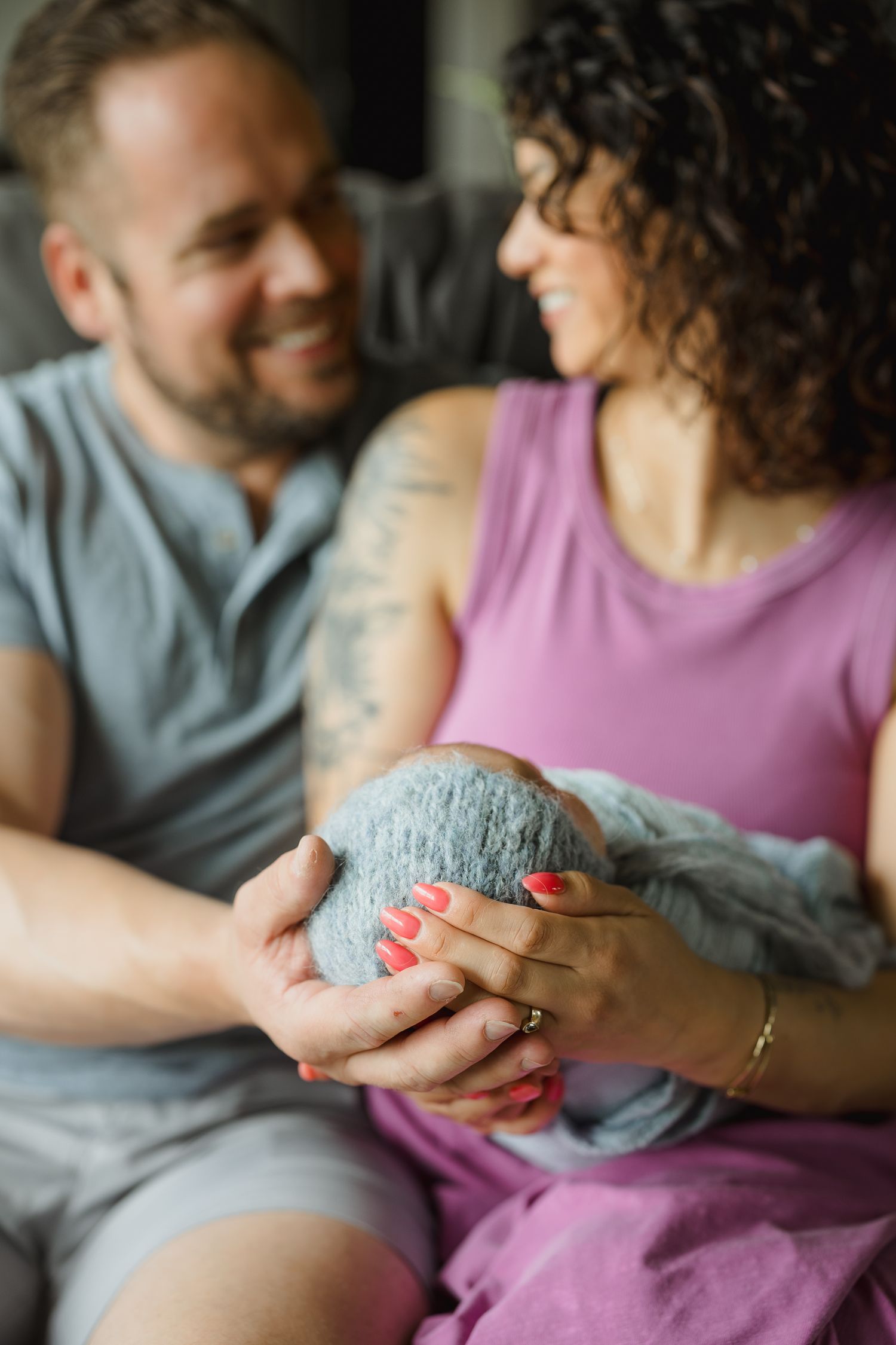 Smiling couple holding their newborn baby together, with the woman in pink and man in gray sitting on a couch.