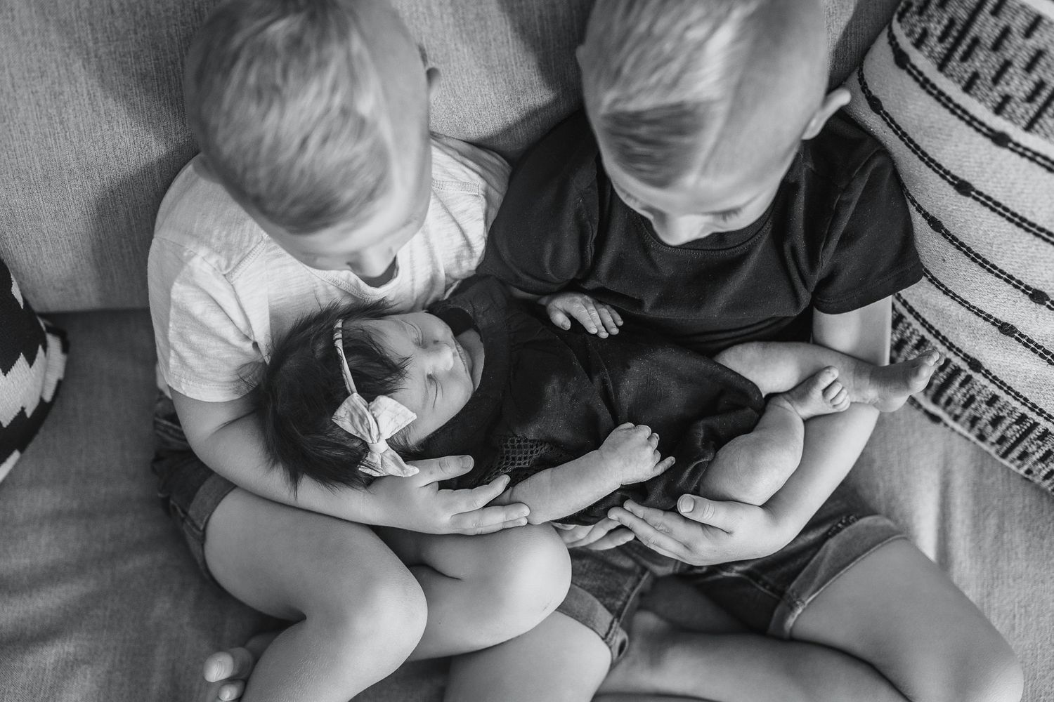 Black and white photo of two young siblings holding a newborn baby on a couch.