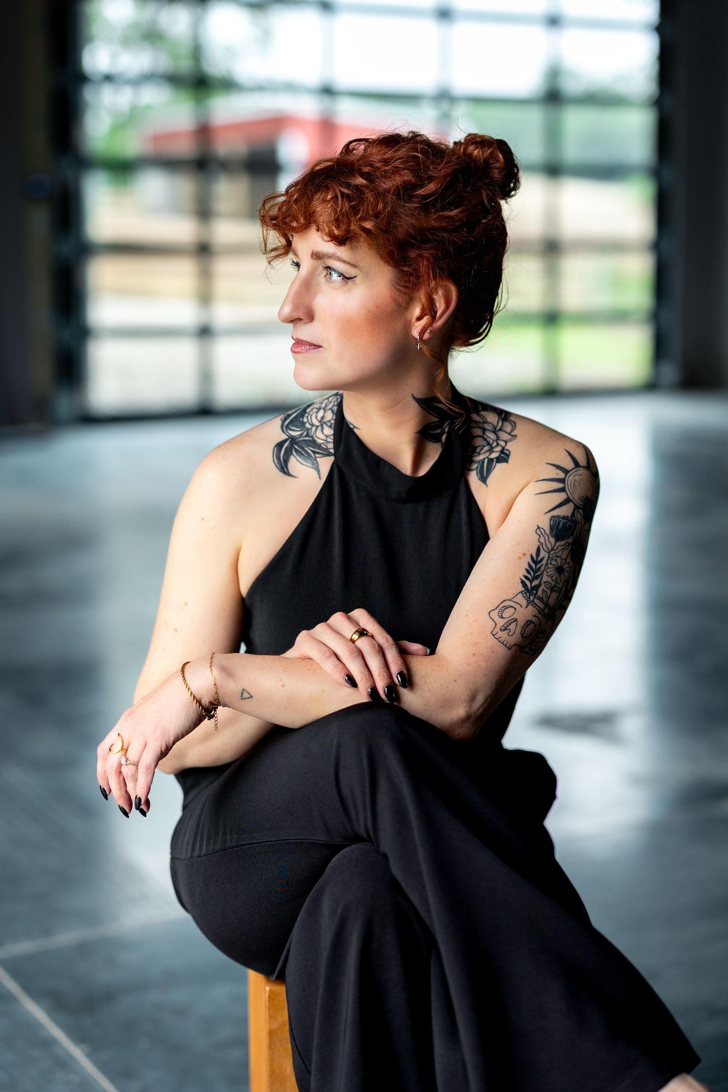 Red-haired woman with tattoos sits gracefully in black halter jumpsuit, gazing sideways in modern studio setting.