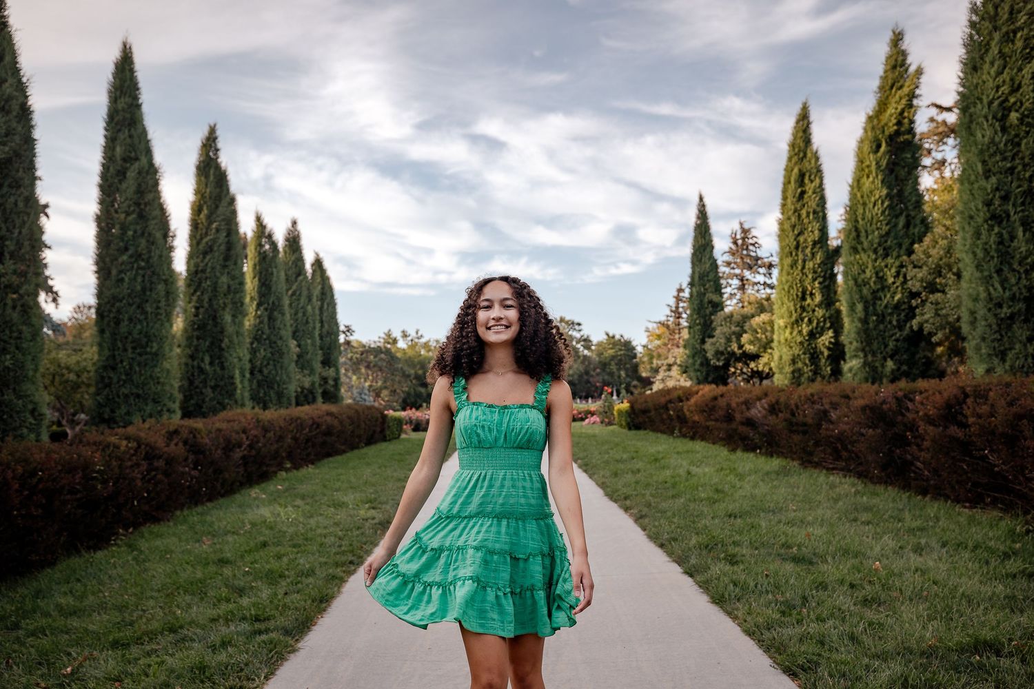 Young woman in green ruffled dress walks along tree-lined garden path under cloudy sky.