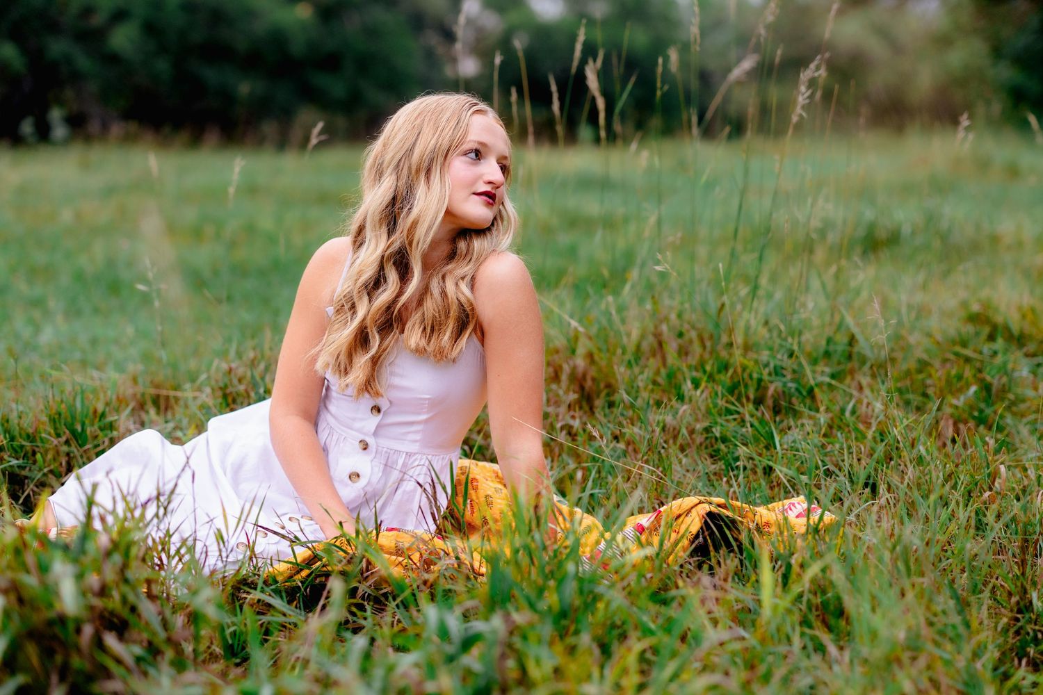Young blonde woman in white dress sitting gracefully in a lush green field, gazing into the distance.