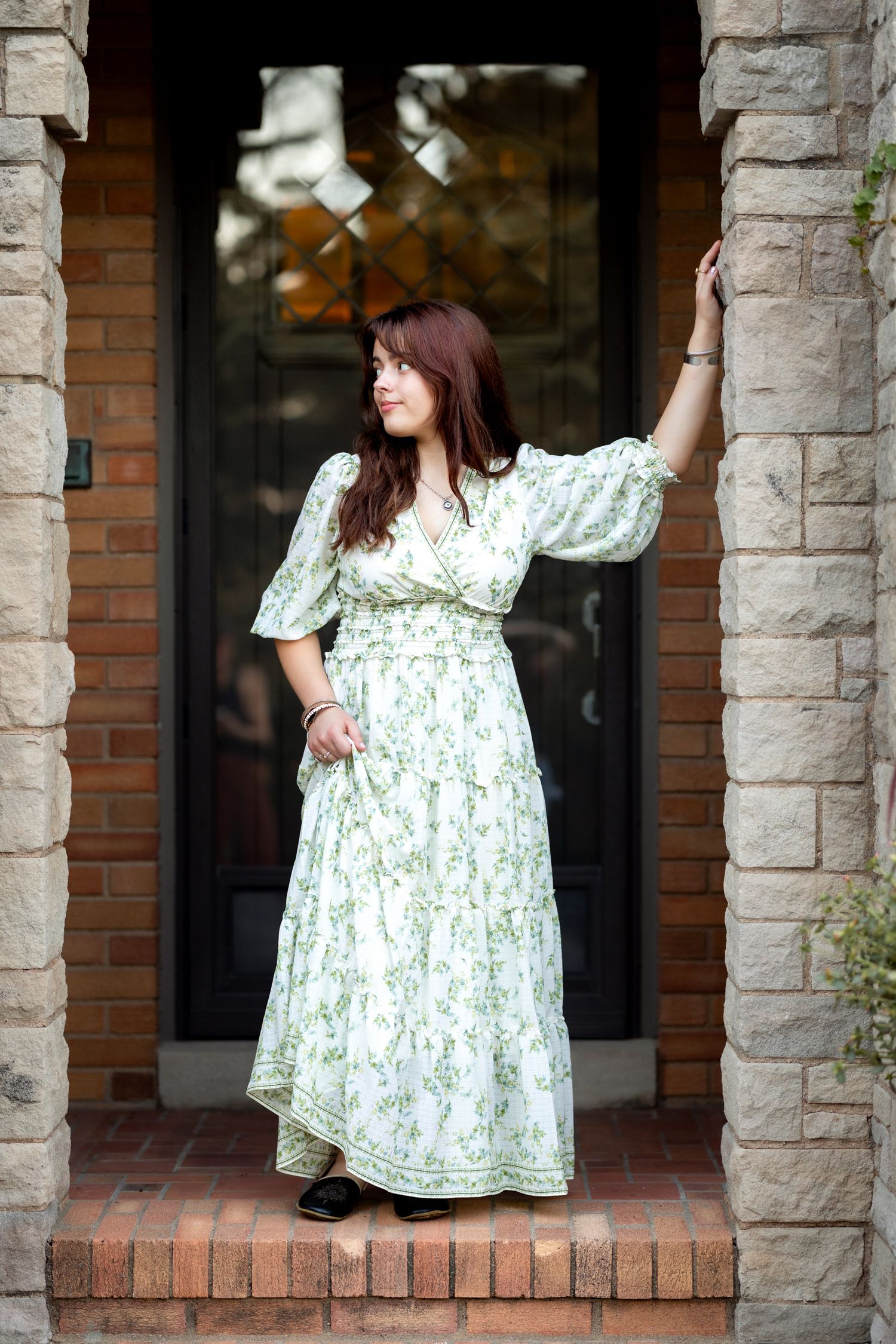 Woman in white floral maxi dress posing on brick steps by ornate door, one arm raised against stone column.