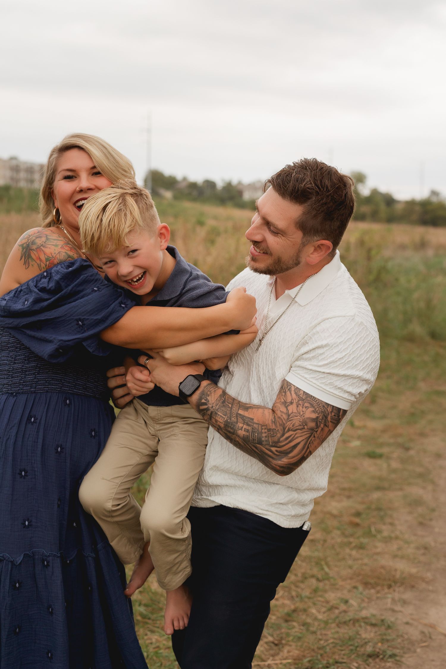 A happy family with tattooed parents playfully lifting their laughing young son outdoors in a field.