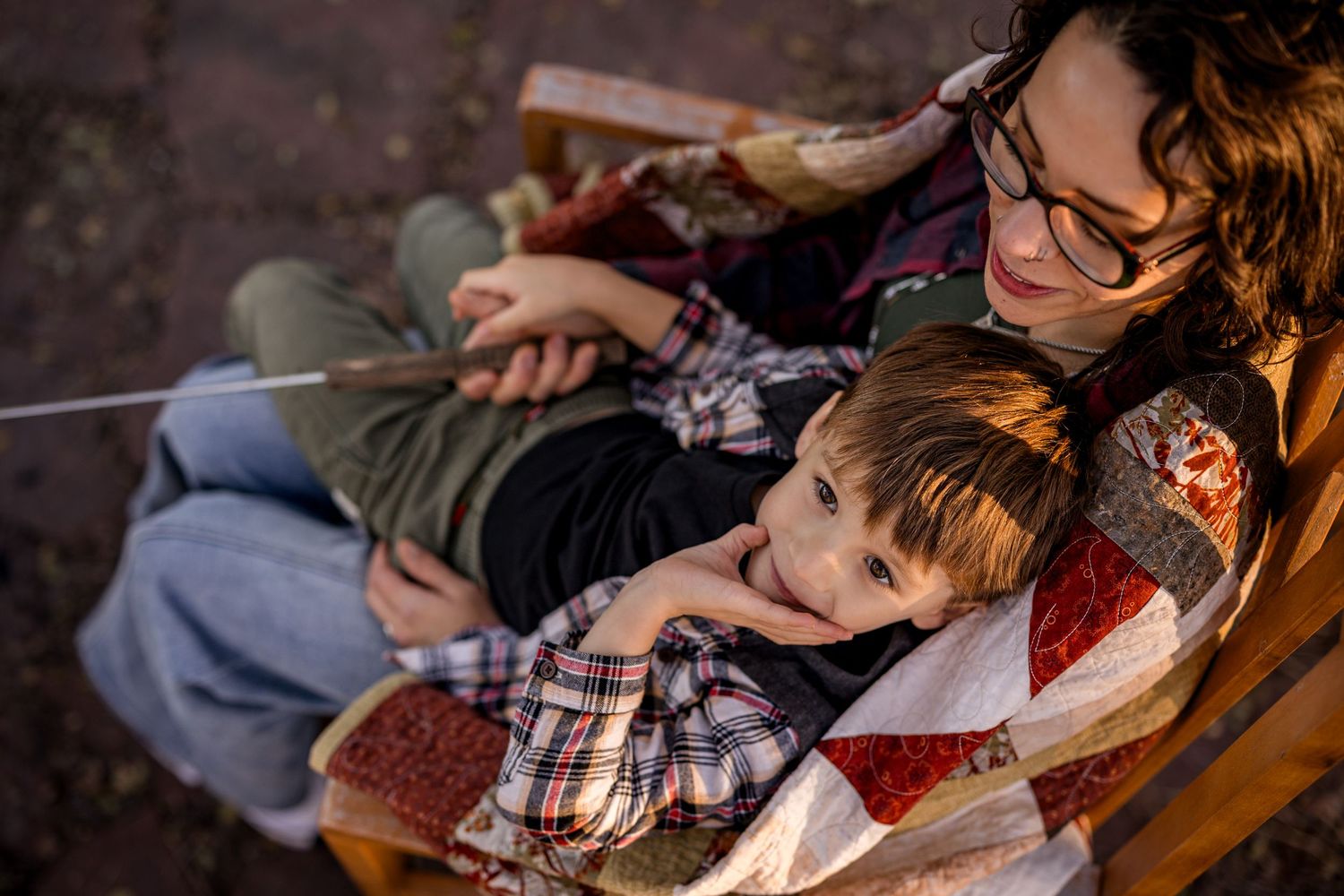 Mother and child snuggled under a quilt on a wooden chair, sharing a warm bonding moment outdoors.