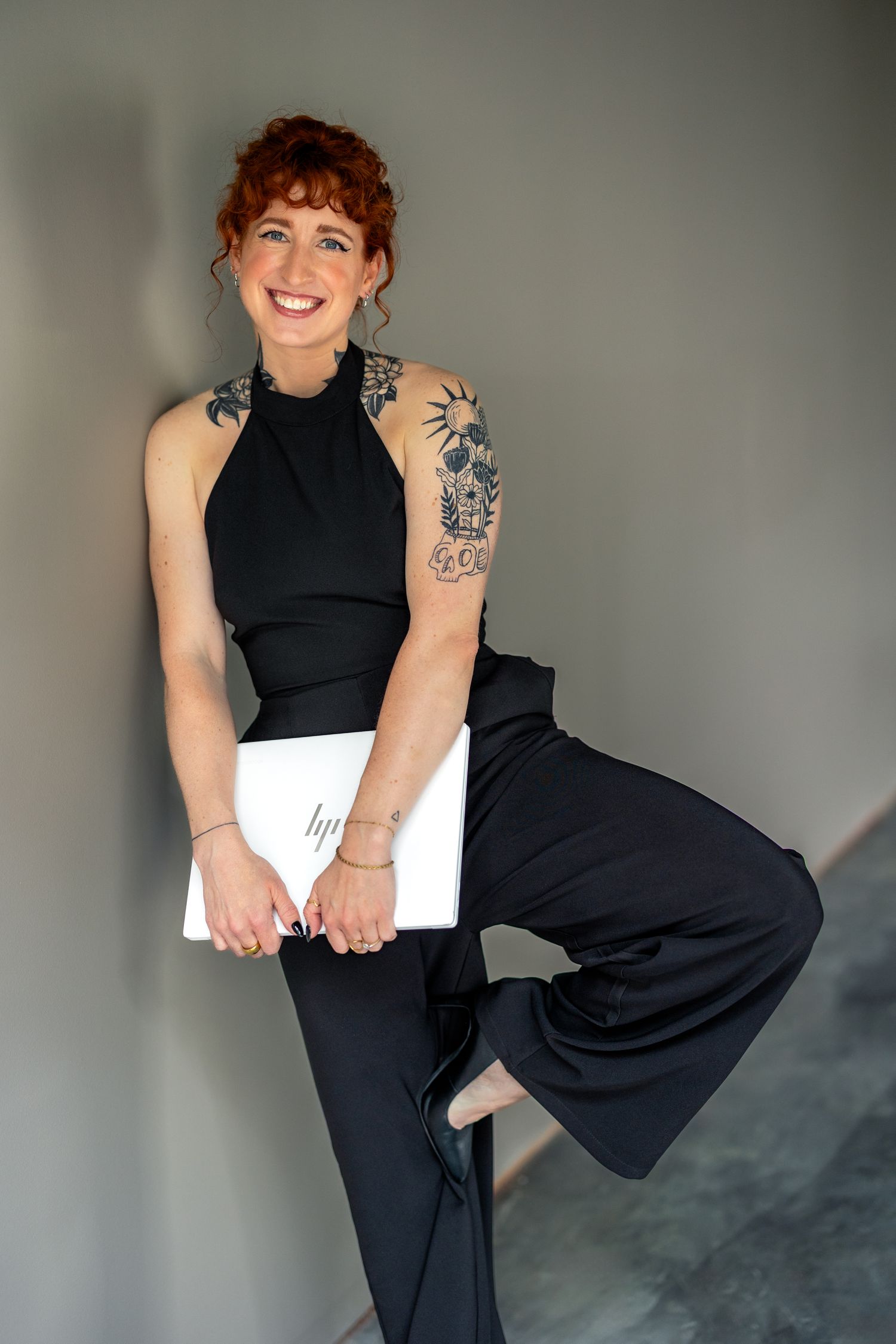 Smiling redhead woman in black halter jumpsuit with tattoos holding an HP laptop, posing confidently against a gray wall.