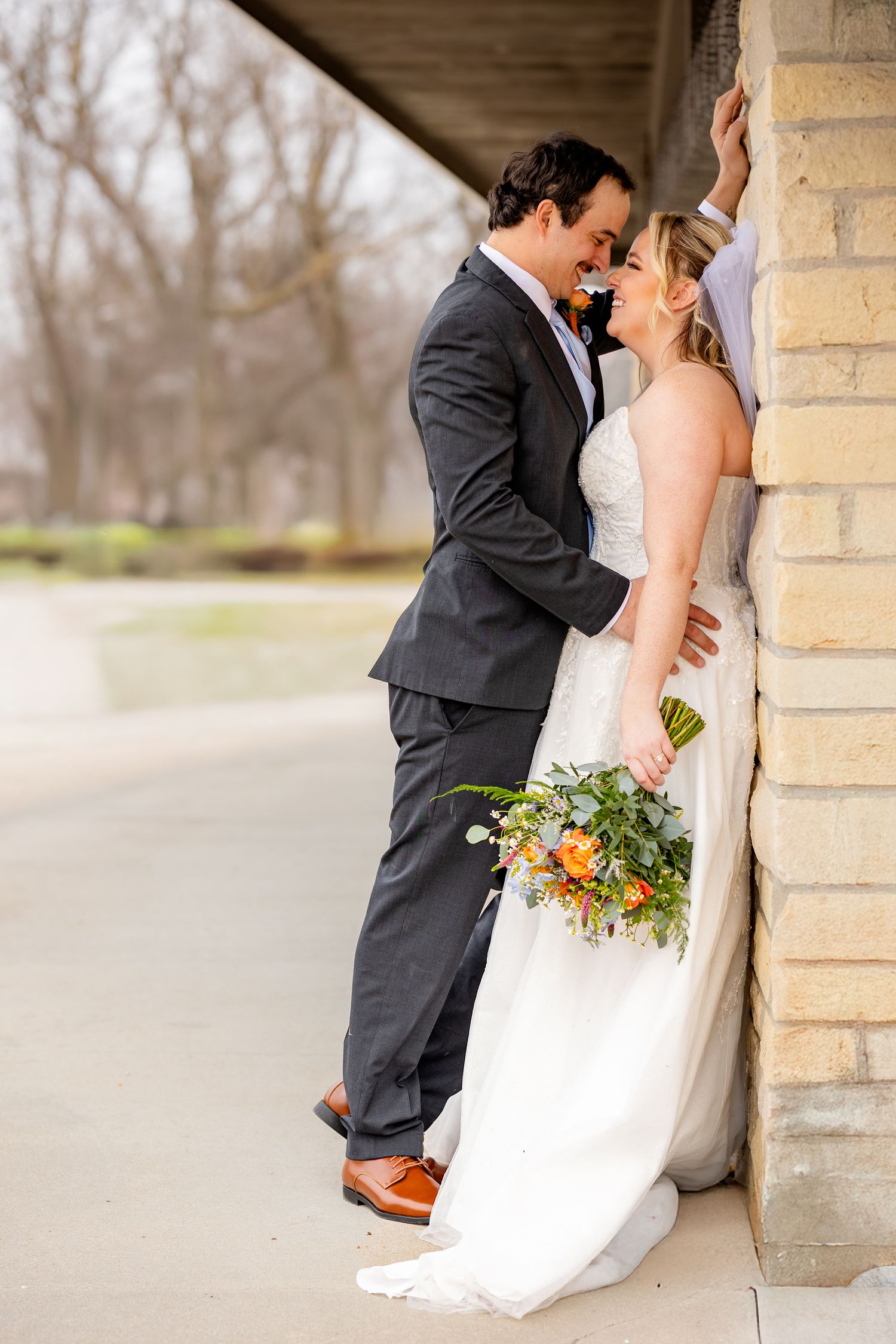 Bride and groom share a romantic moment leaning against a brick wall, bride holding a colorful orange and green bouquet.