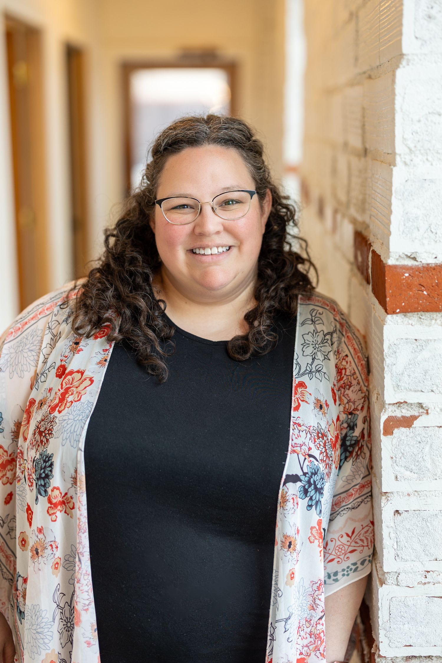 Smiling woman with curly hair and glasses wearing a floral cardigan, leaning against a white brick wall in a hallway.