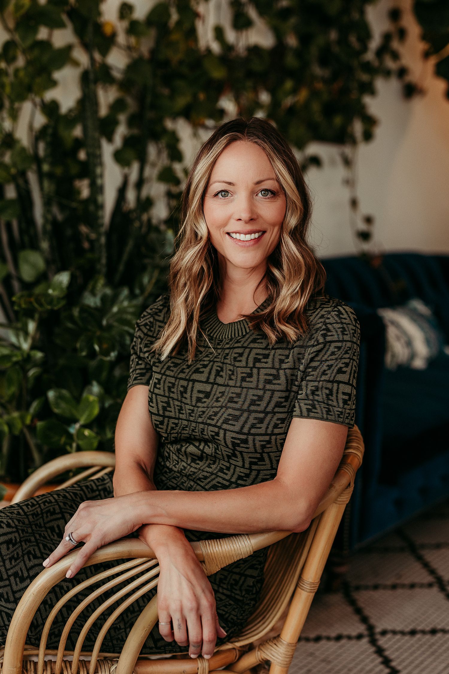 Smiling woman with blonde hair sits in rattan chair wearing patterned dress, surrounded by lush green plants indoors.