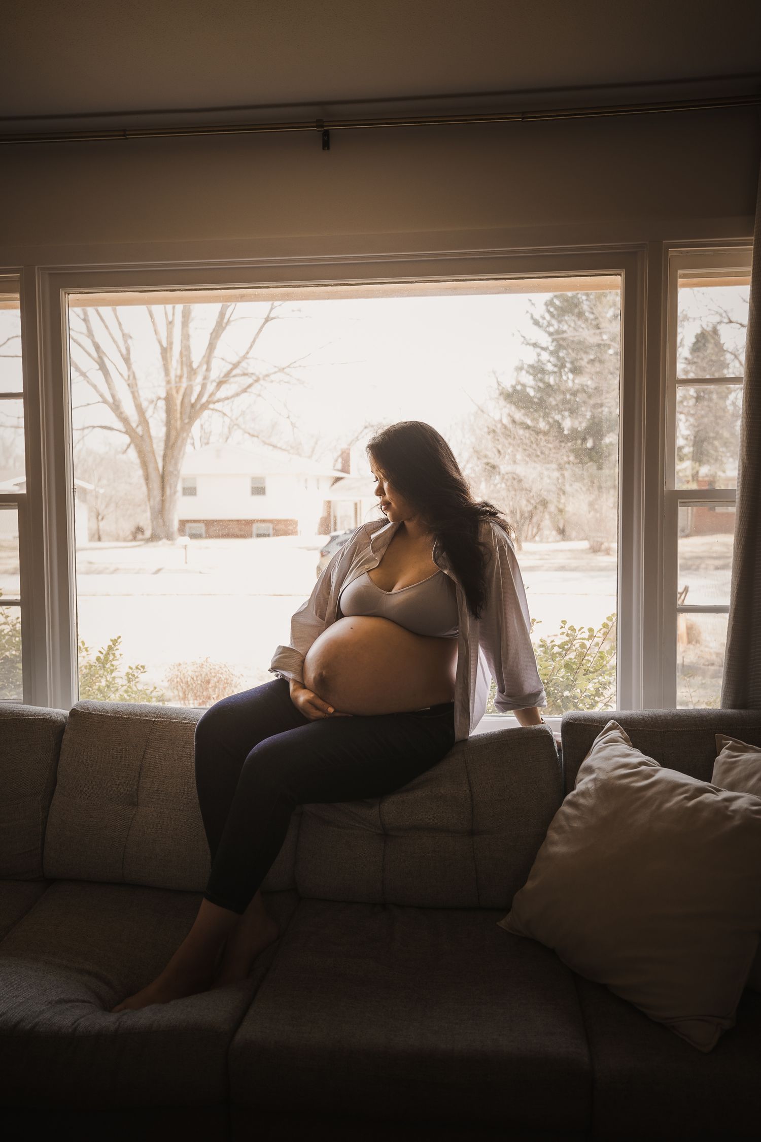 Pregnant woman sitting on couch by large window, cradling baby bump in soft natural light.