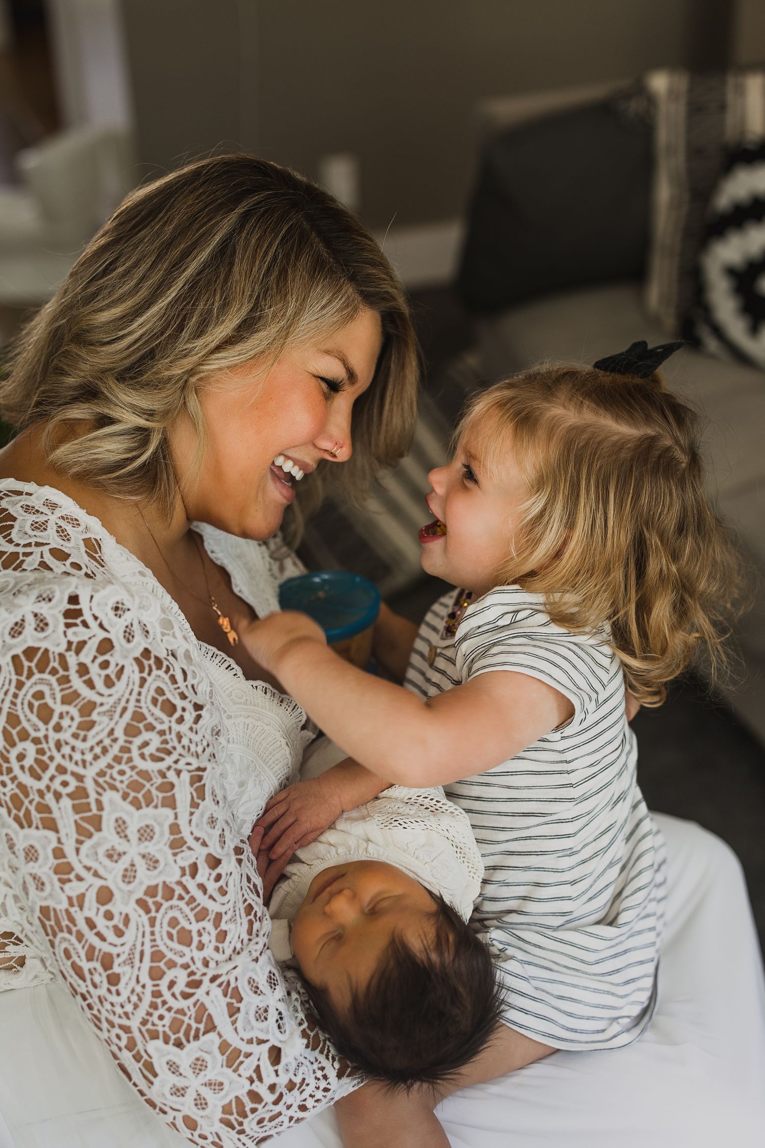 Smiling mother in white lace dress holds newborn while toddler girl in striped shirt reaches out joyfully.