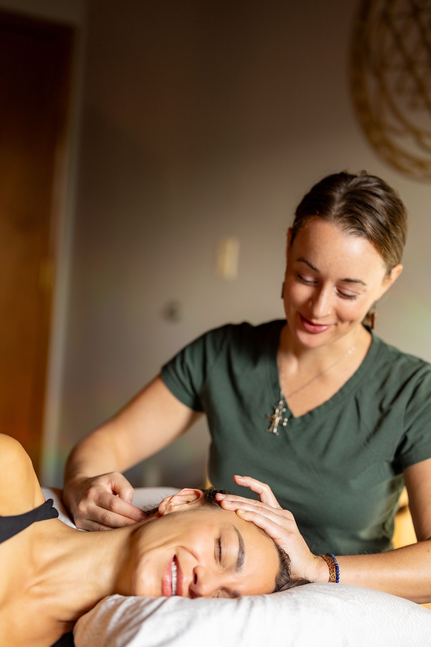 Smiling massage therapist in green scrubs performing a relaxing facial massage on a client lying on a treatment table.