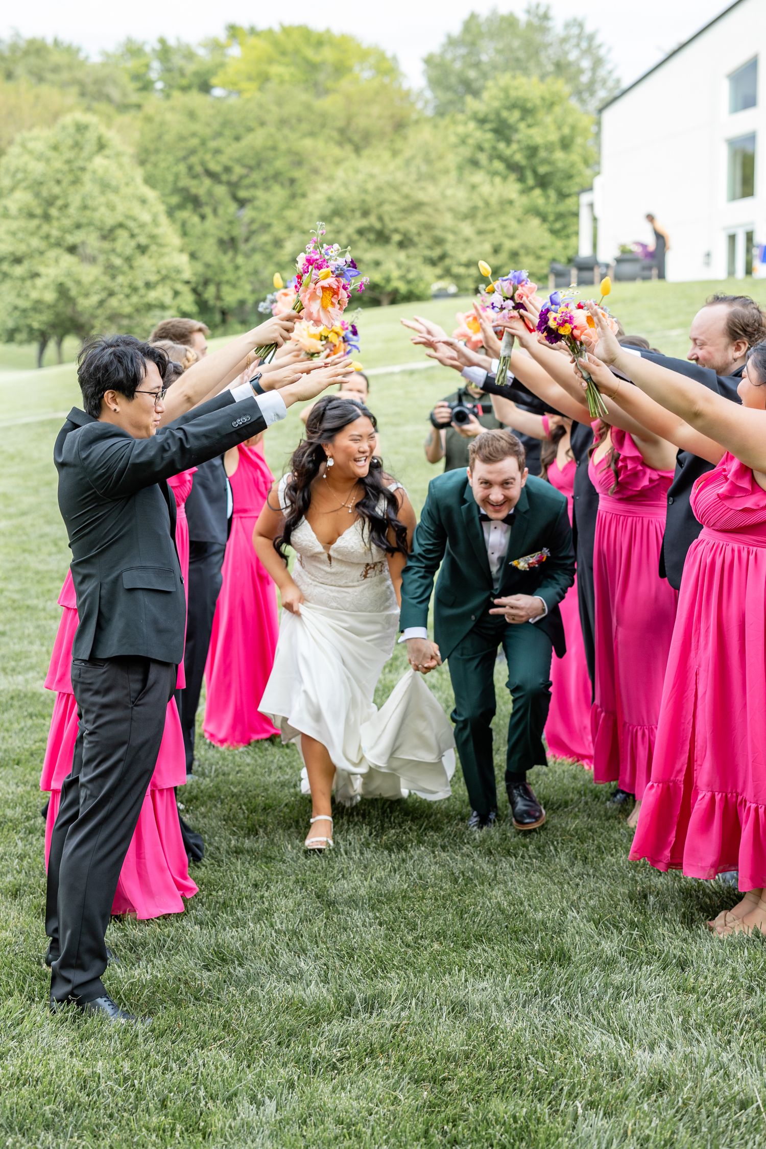 Bride and groom run through a tunnel of cheering wedding party in pink dresses on a green lawn.