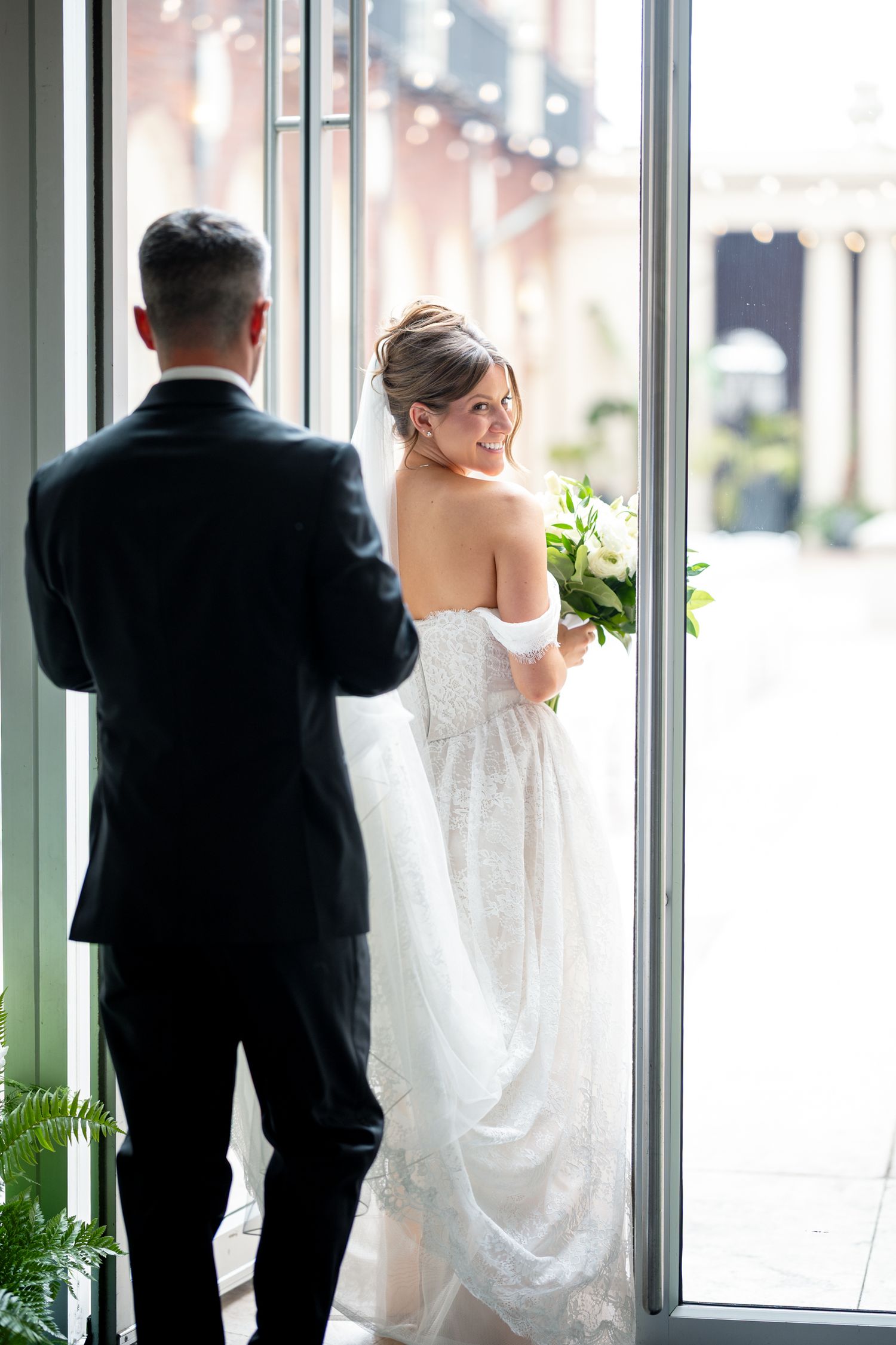 Bride in white off-shoulder gown holds bouquet, smiling back at groom in black suit near large windows.