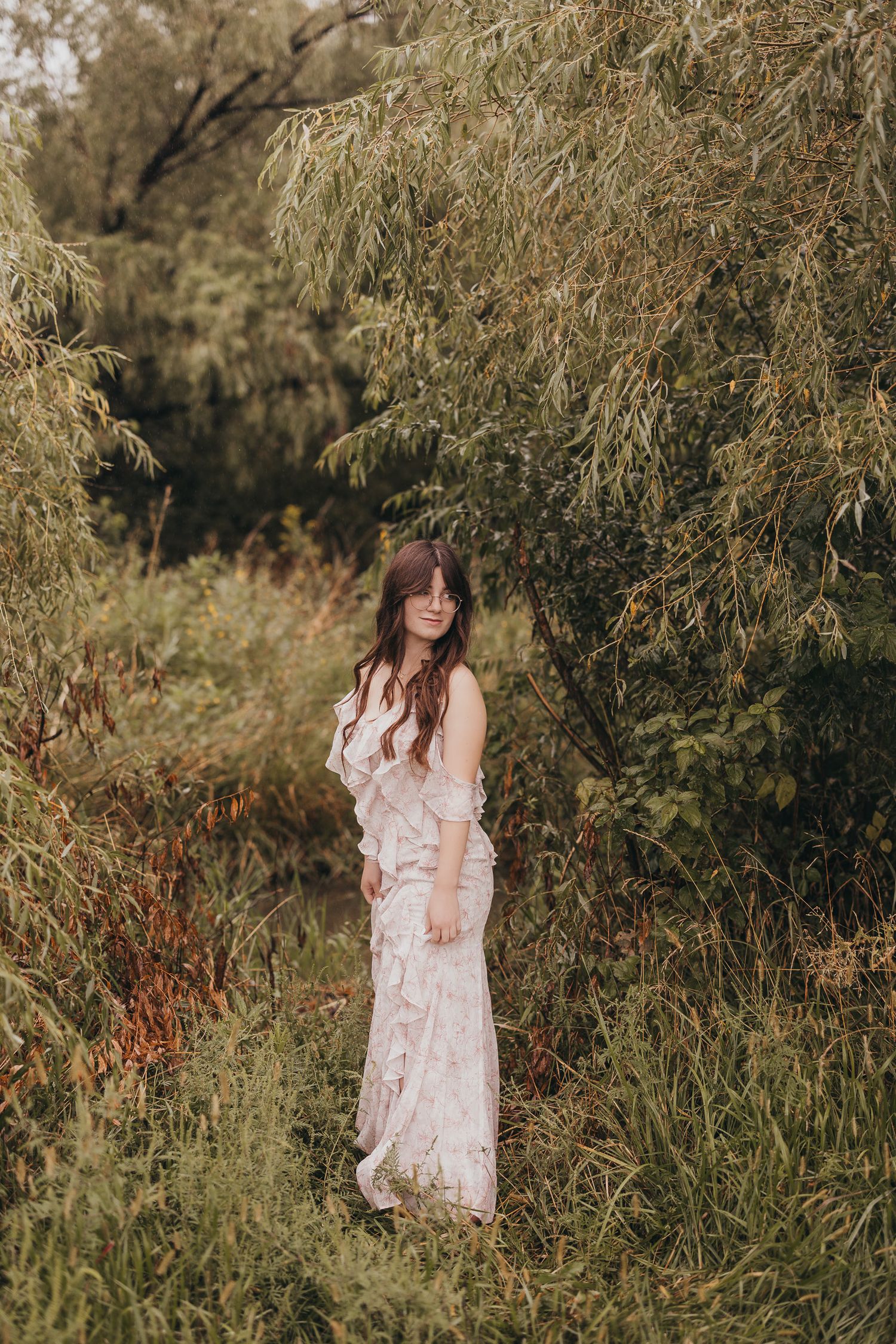 Figure in a flowy white lace dress poses among tall grasses and trees in a dreamy outdoor setting.