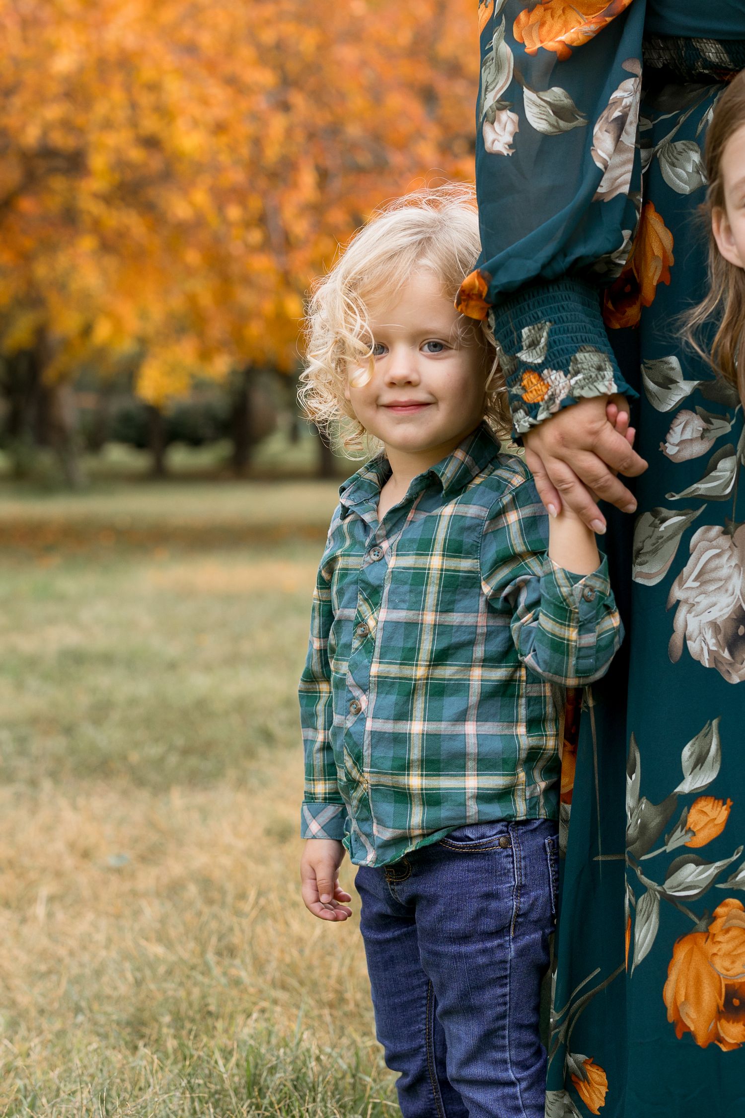 Curly-haired toddler in plaid shirt stands beside adult in floral dress among autumn trees.
