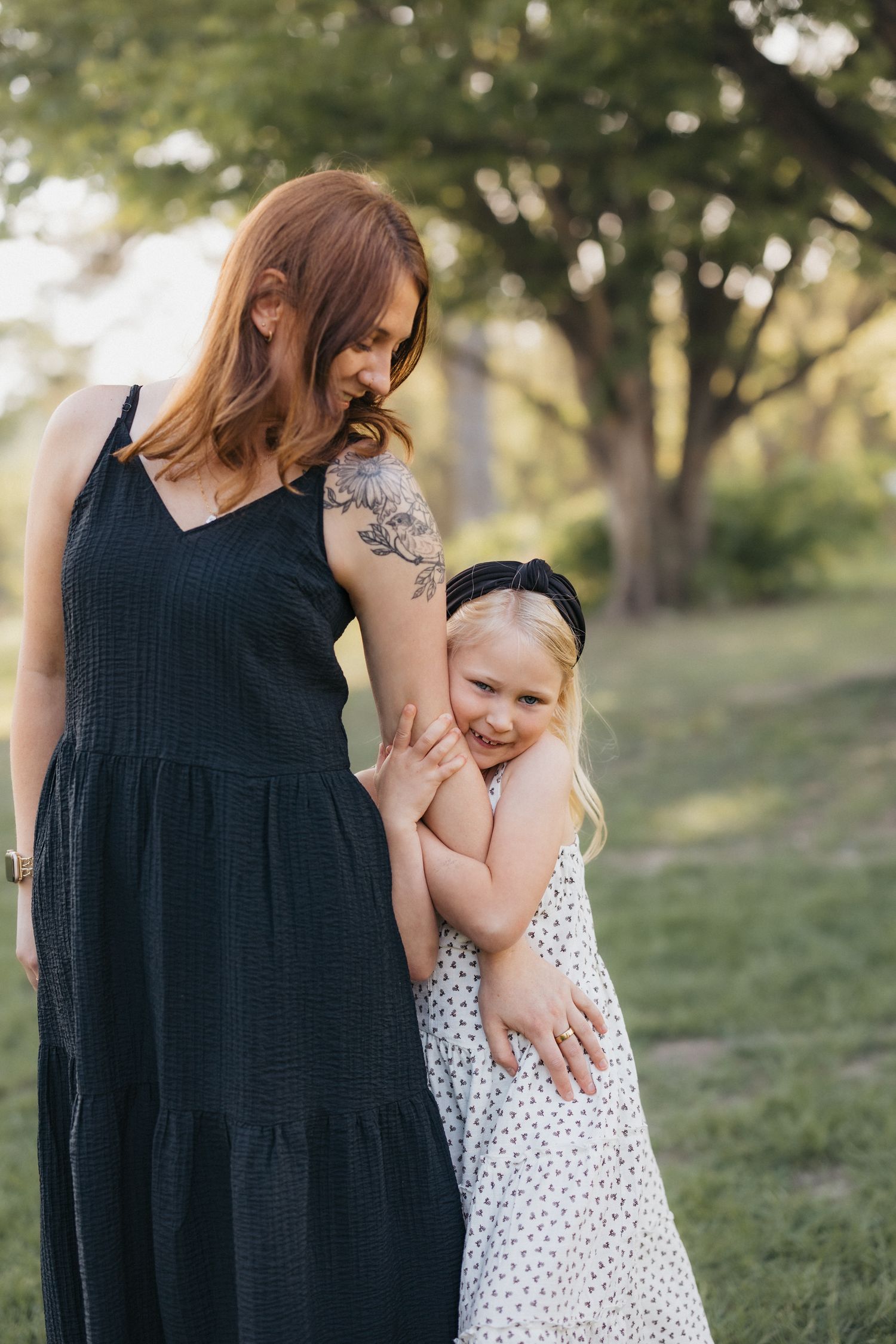 A redhead mother with a tattoo in a black dress hugs her smiling blonde daughter in a white polka dot dress outdoors.