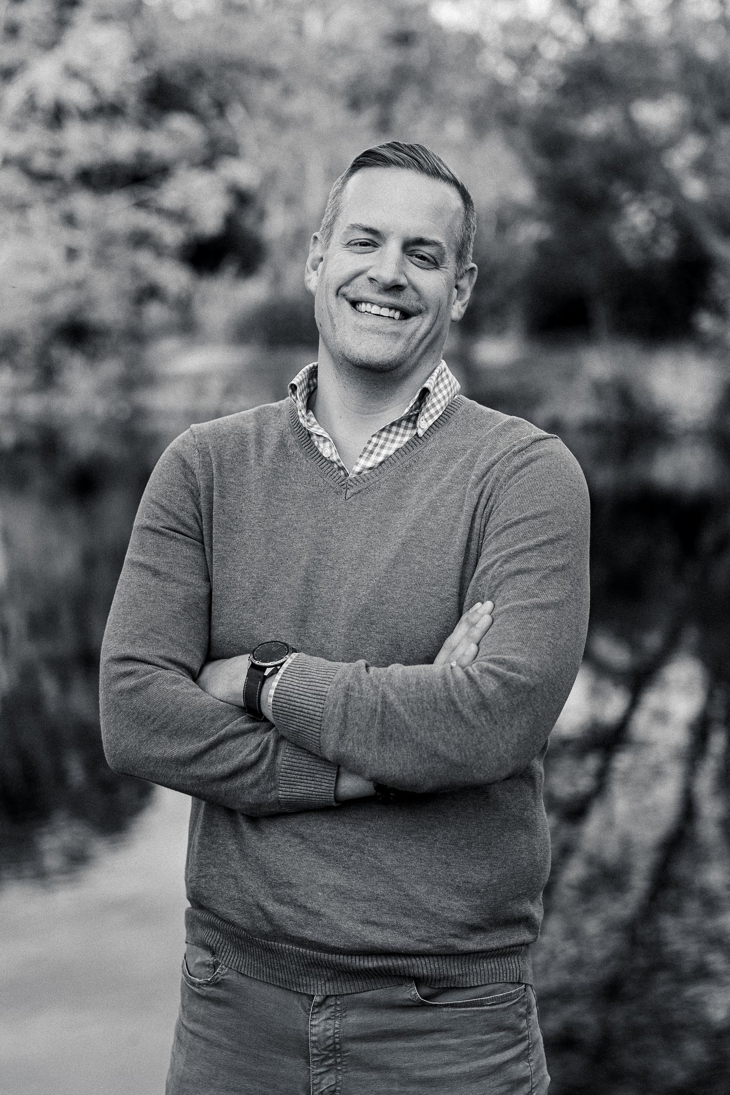 Smiling man with arms crossed poses outdoors near a pond in a black and white portrait photo.