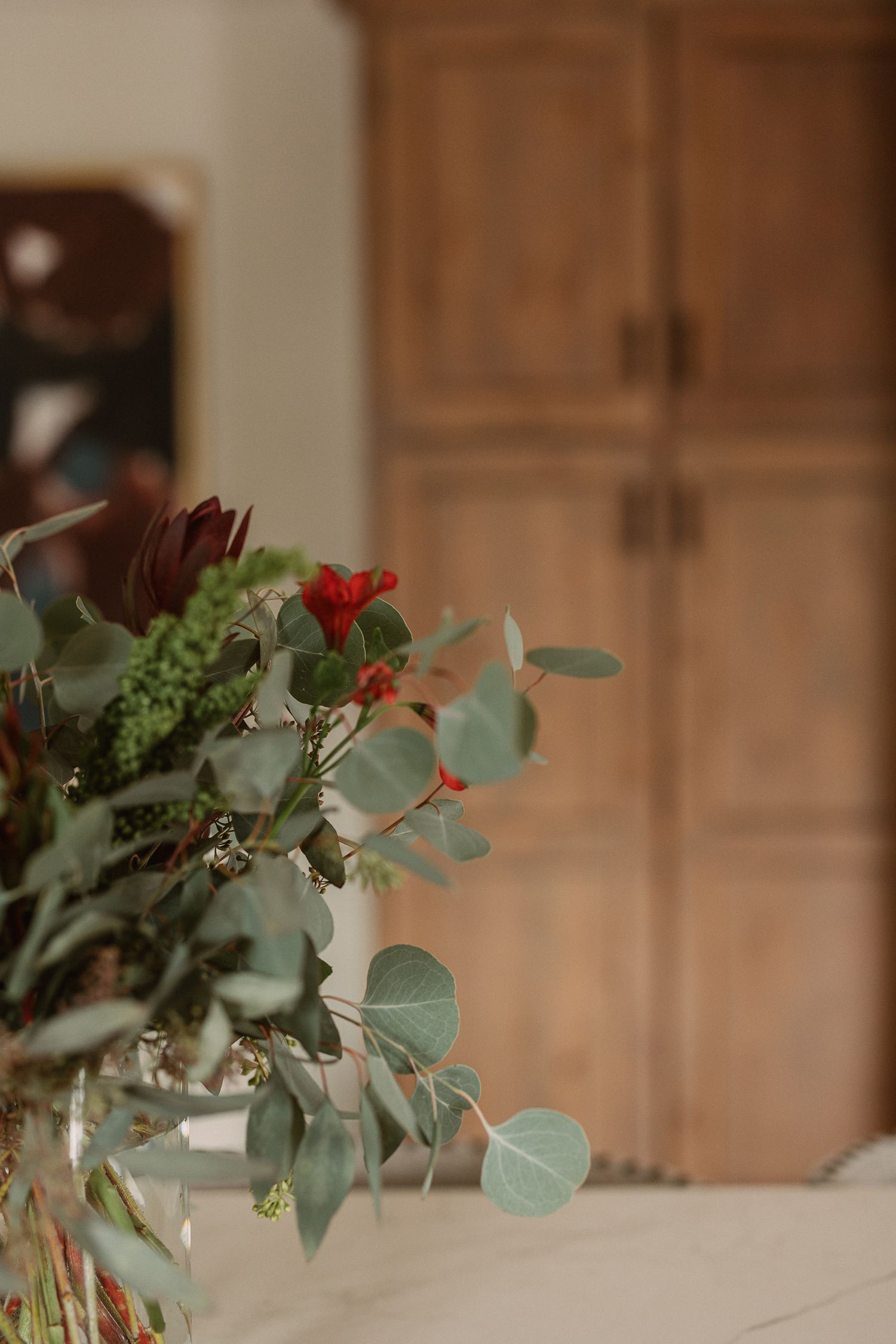 Lush eucalyptus and red flower bouquet arrangement on a white surface with warm wooden cabinet in the background.