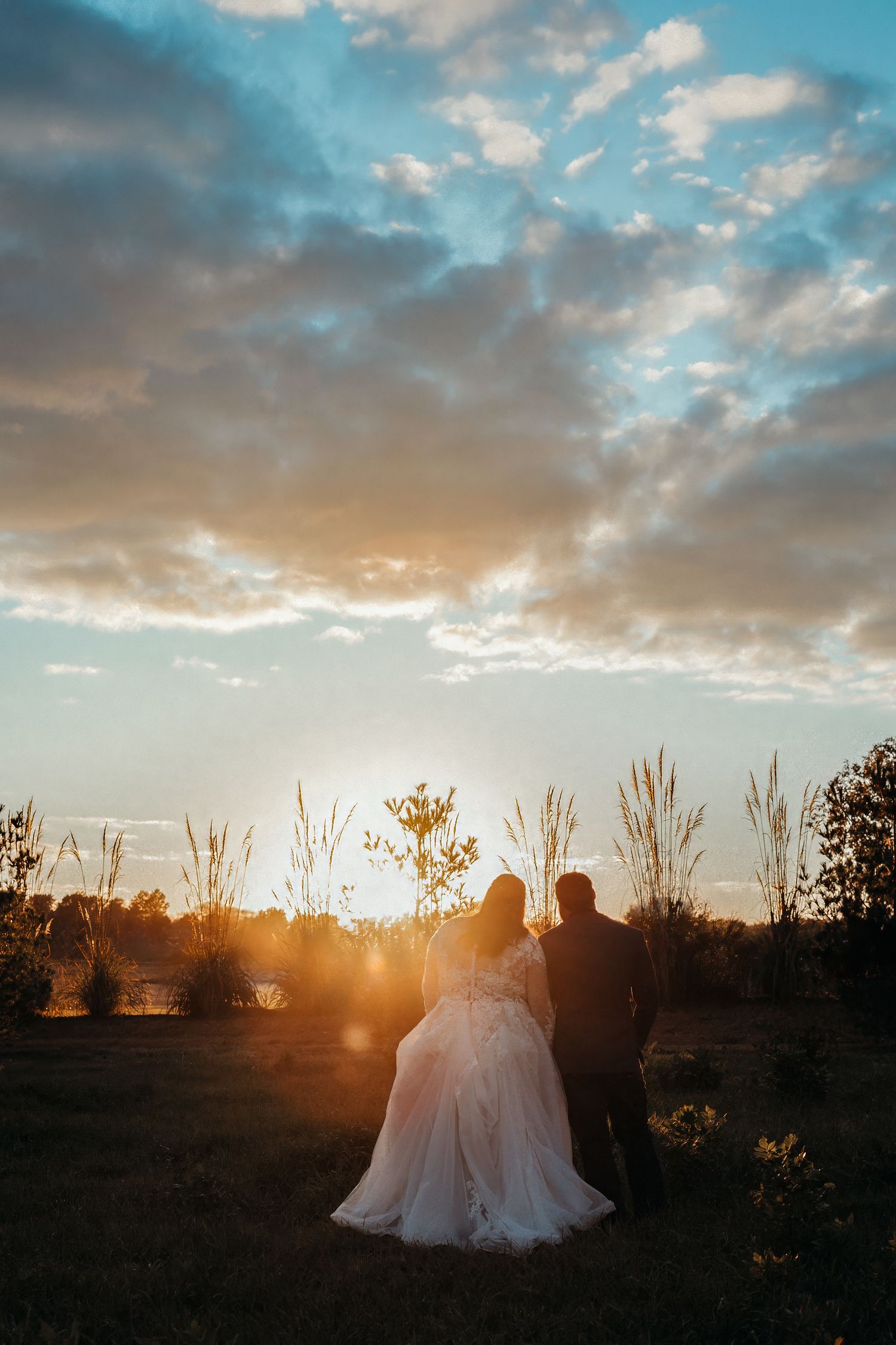 Bride and groom silhouetted against a golden sunset with dramatic clouds in an outdoor wedding portrait.