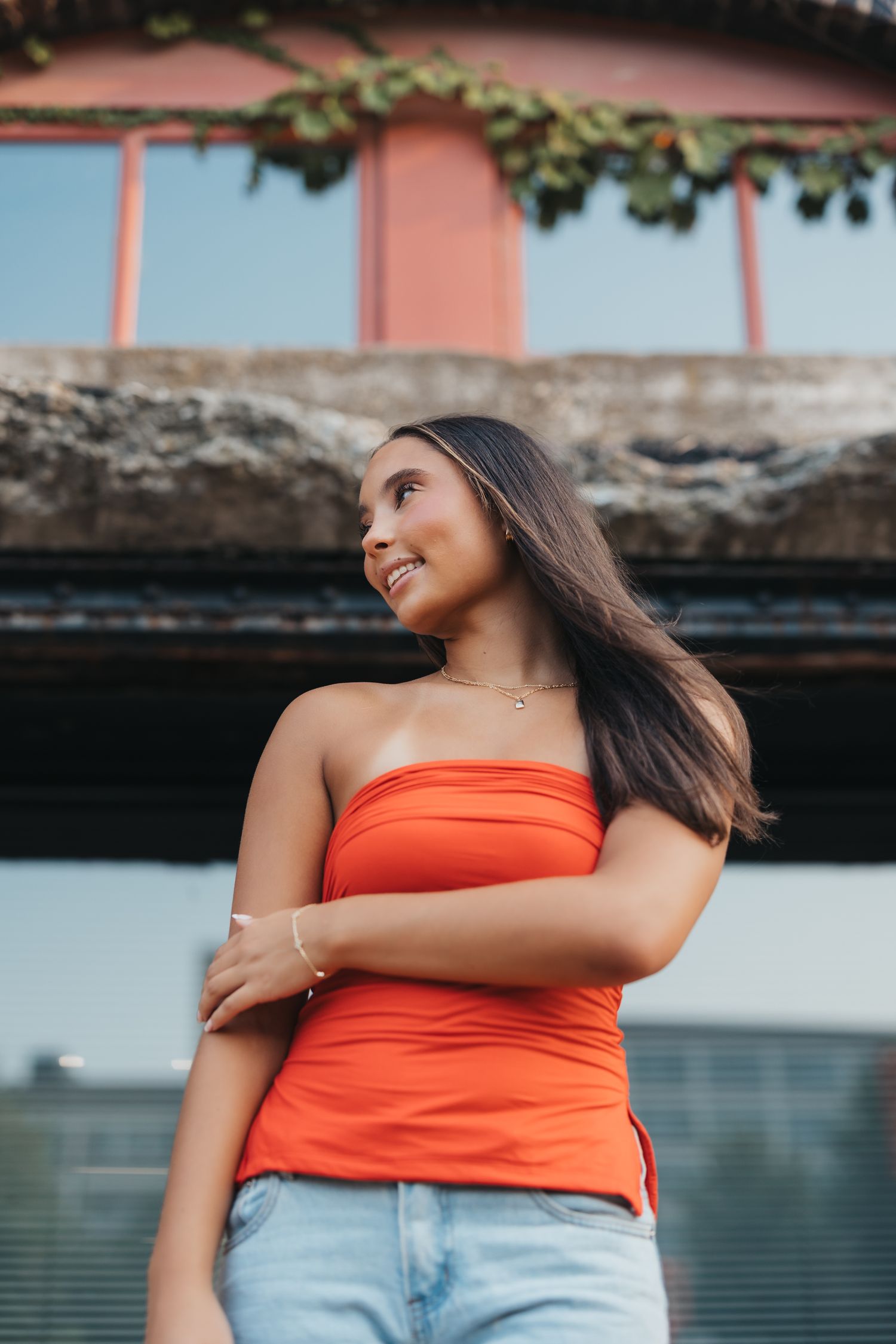 Young woman in orange tube top and jeans smiling outdoors near a bridge on a sunny day.