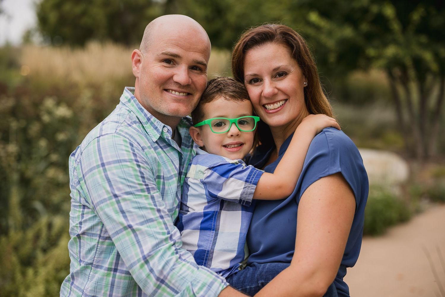 Happy family portrait with parents and young boy wearing green glasses smiling outdoors in a garden setting.