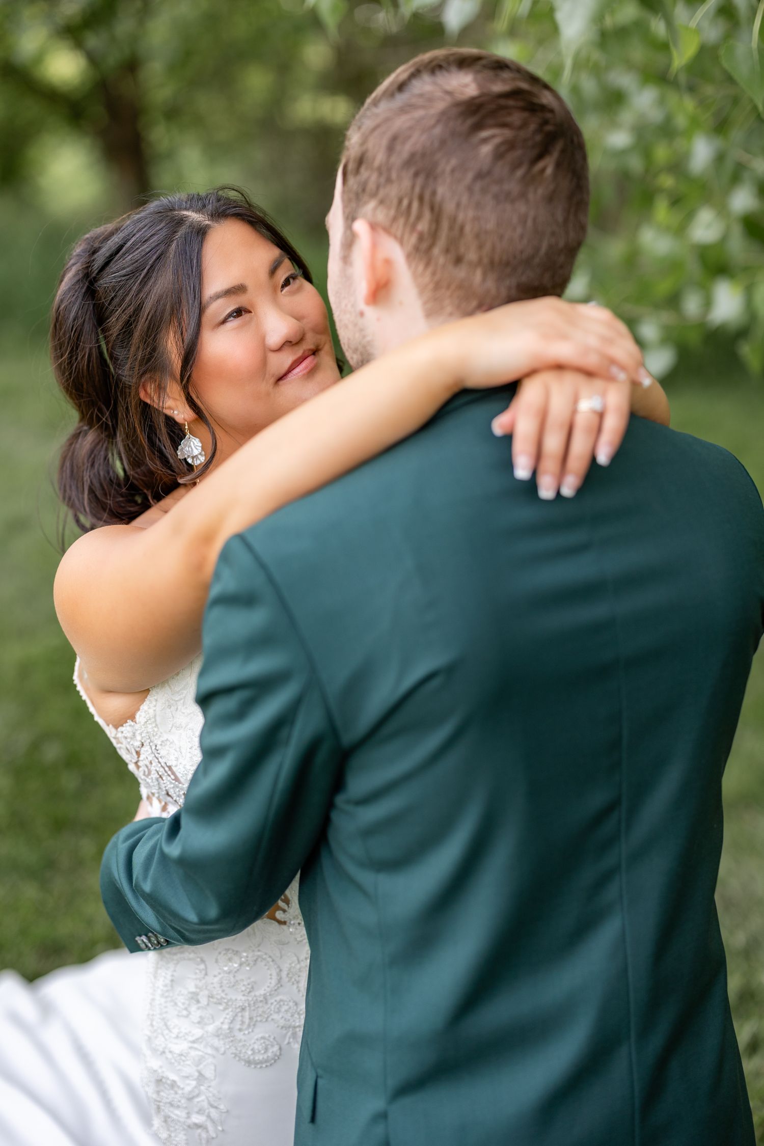 Bride in lace gown embraces groom in green suit outdoors, smiling warmly, showcasing her diamond engagement ring.