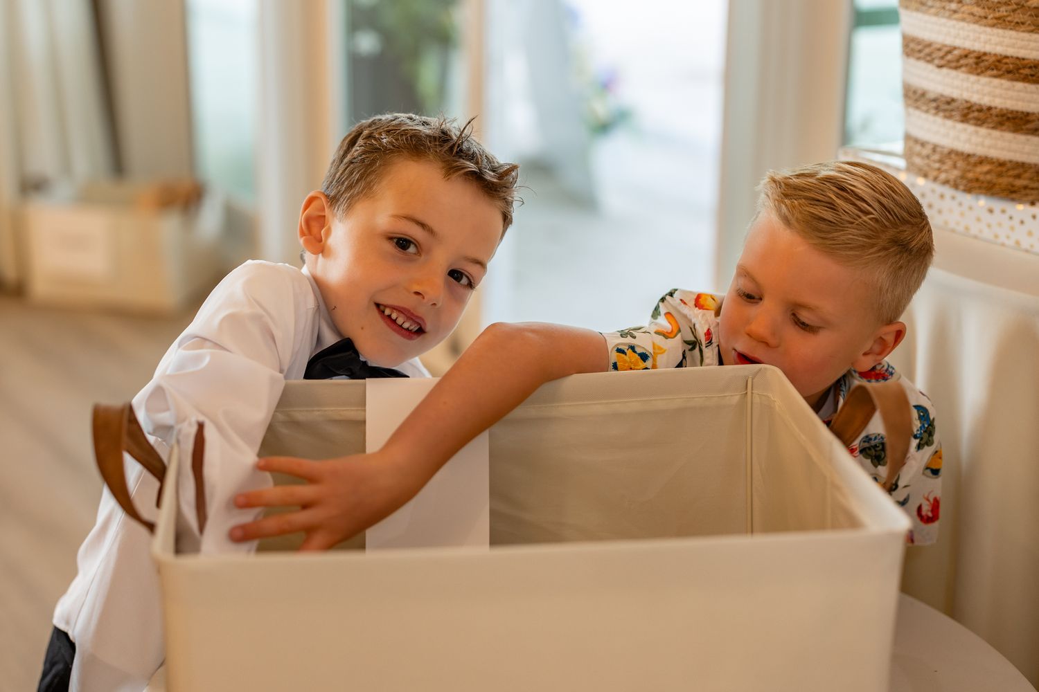 Two young boys smiling and playing together with a large white storage box indoors.
