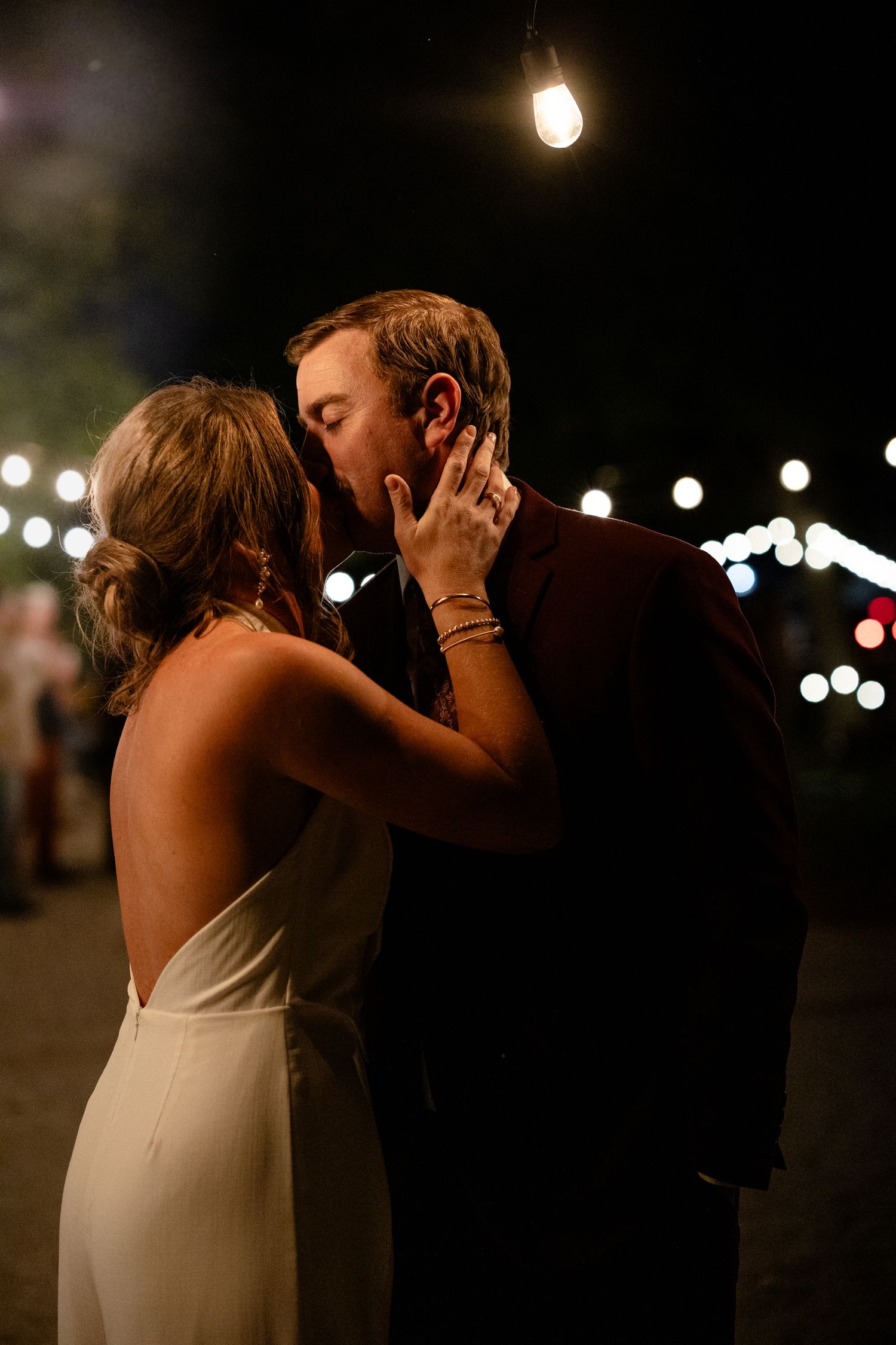 Bride and groom share a romantic first kiss at night, surrounded by warm string lights at their wedding reception.