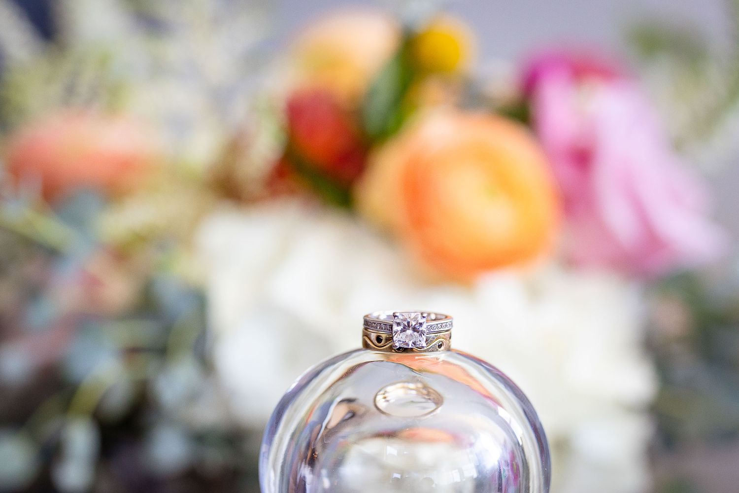 Close-up of diamond engagement ring and wedding band balanced on glass with colorful floral bouquet background.