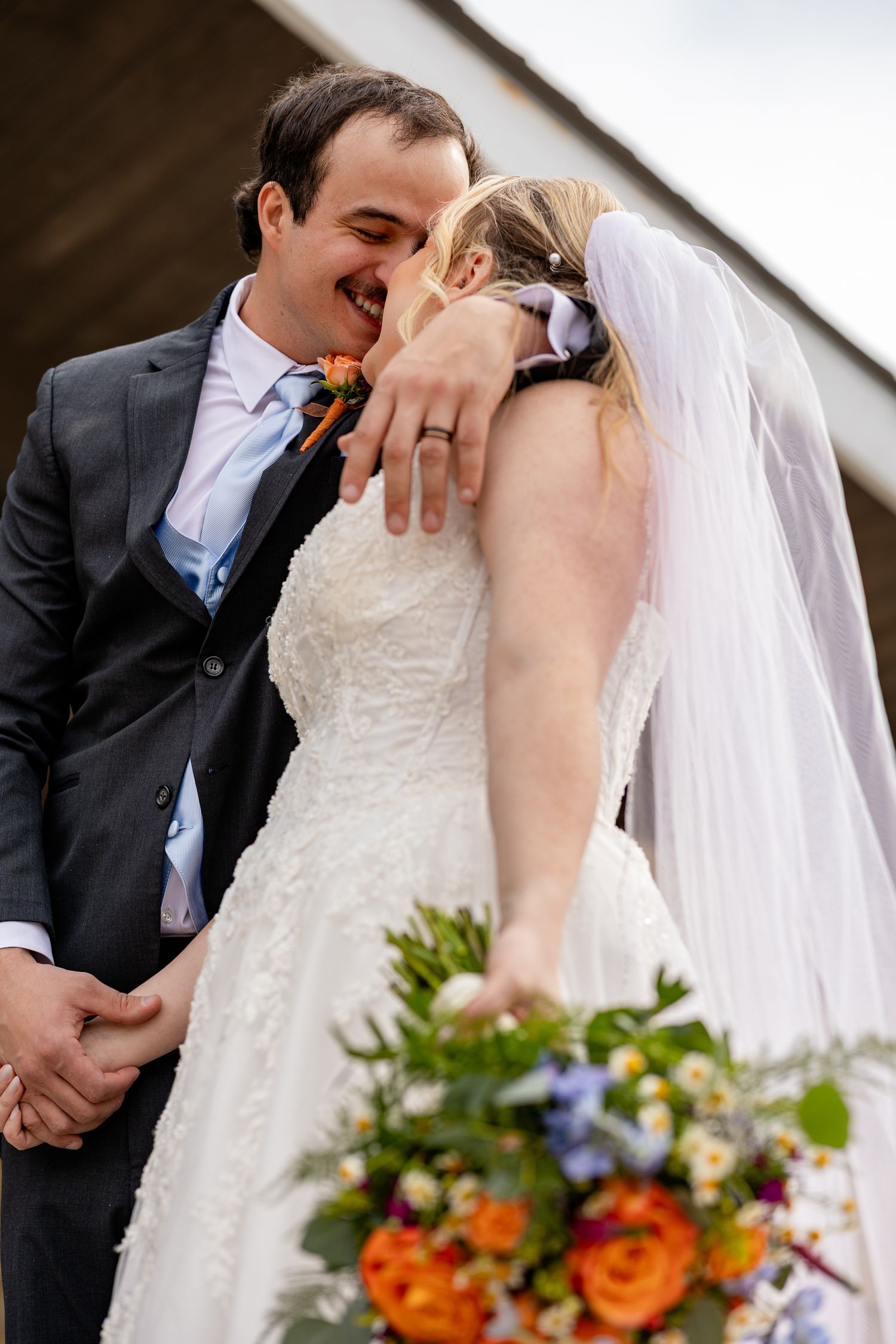 Bride and groom share a joyful kiss, bride holds colorful bouquet of orange and white flowers, wearing white lace gown and veil.