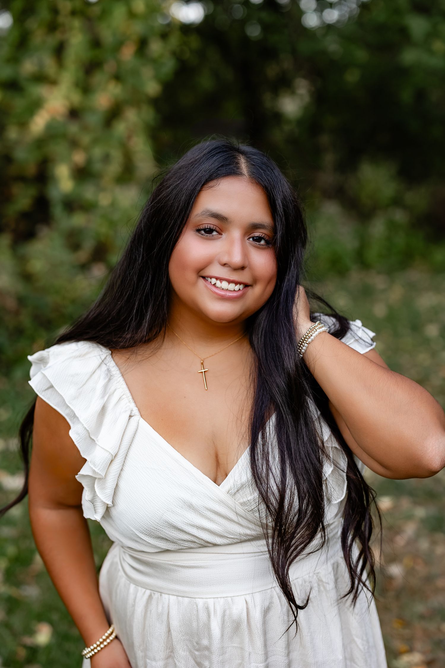 Young woman in white ruffled dress with gold cross necklace smiling outdoors among green trees.