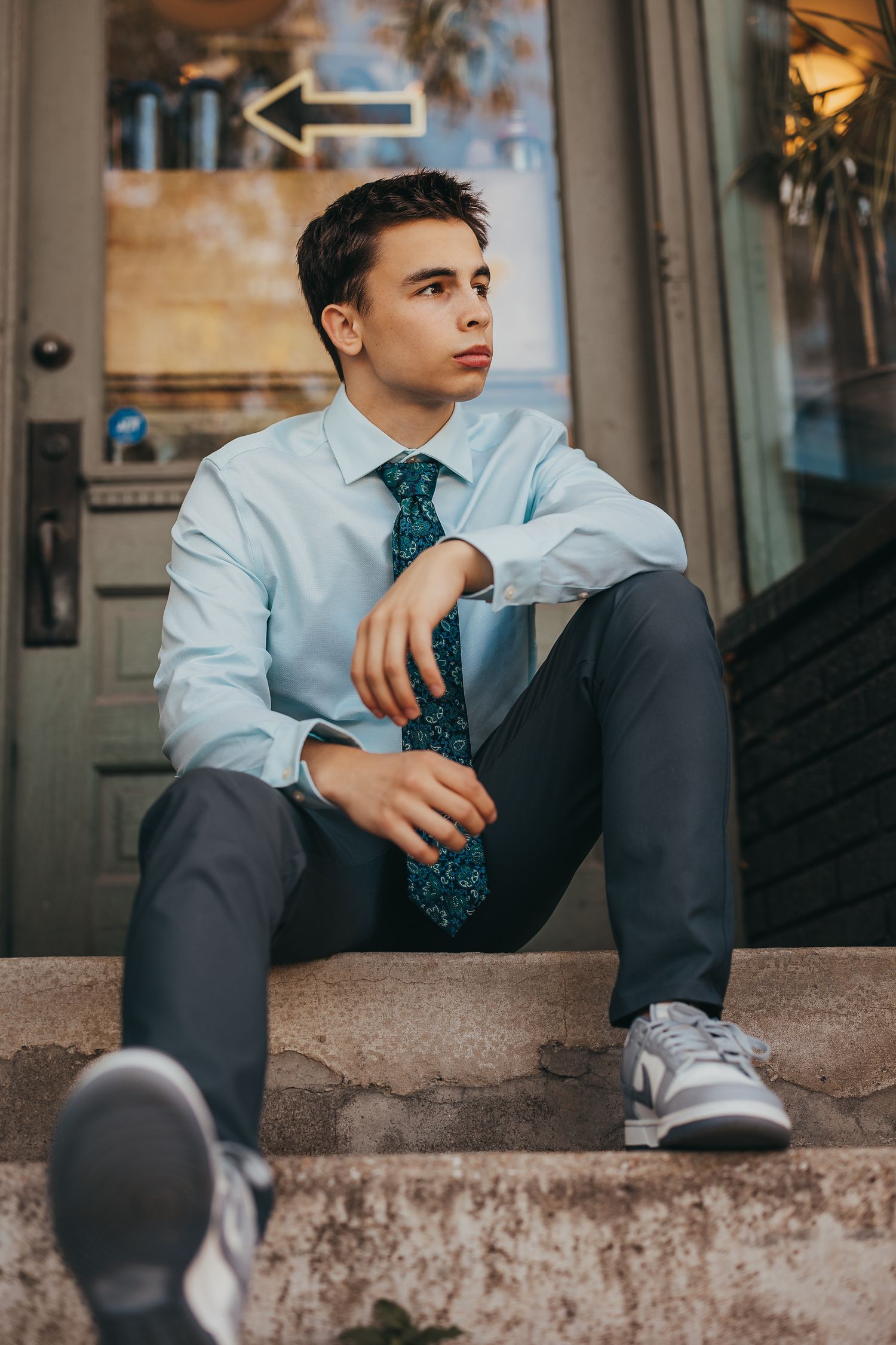 Young man in light blue dress shirt and teal tie sitting on stone steps outdoors, looking thoughtfully to the side.