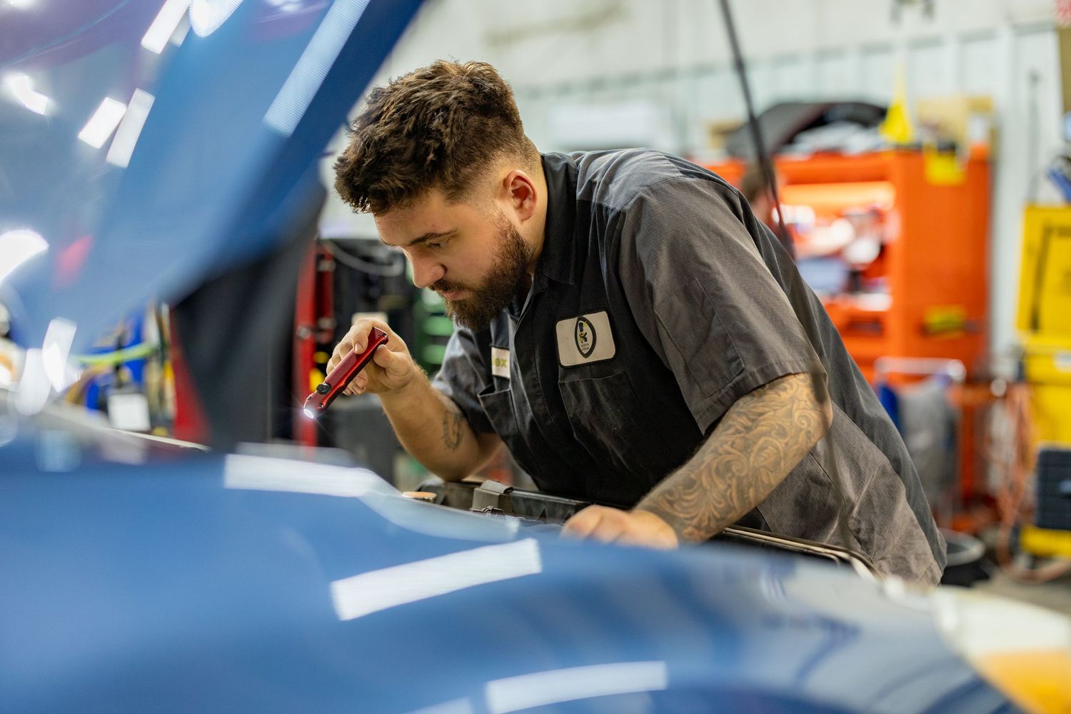 A mechanic inspects a blue car engine in a professional auto repair shop, wearing a gray uniform.