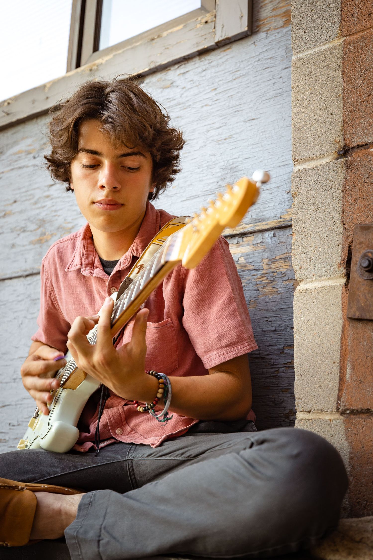 A teenage boy with curly hair sits cross-legged playing an electric guitar against a rustic brick and wood wall.