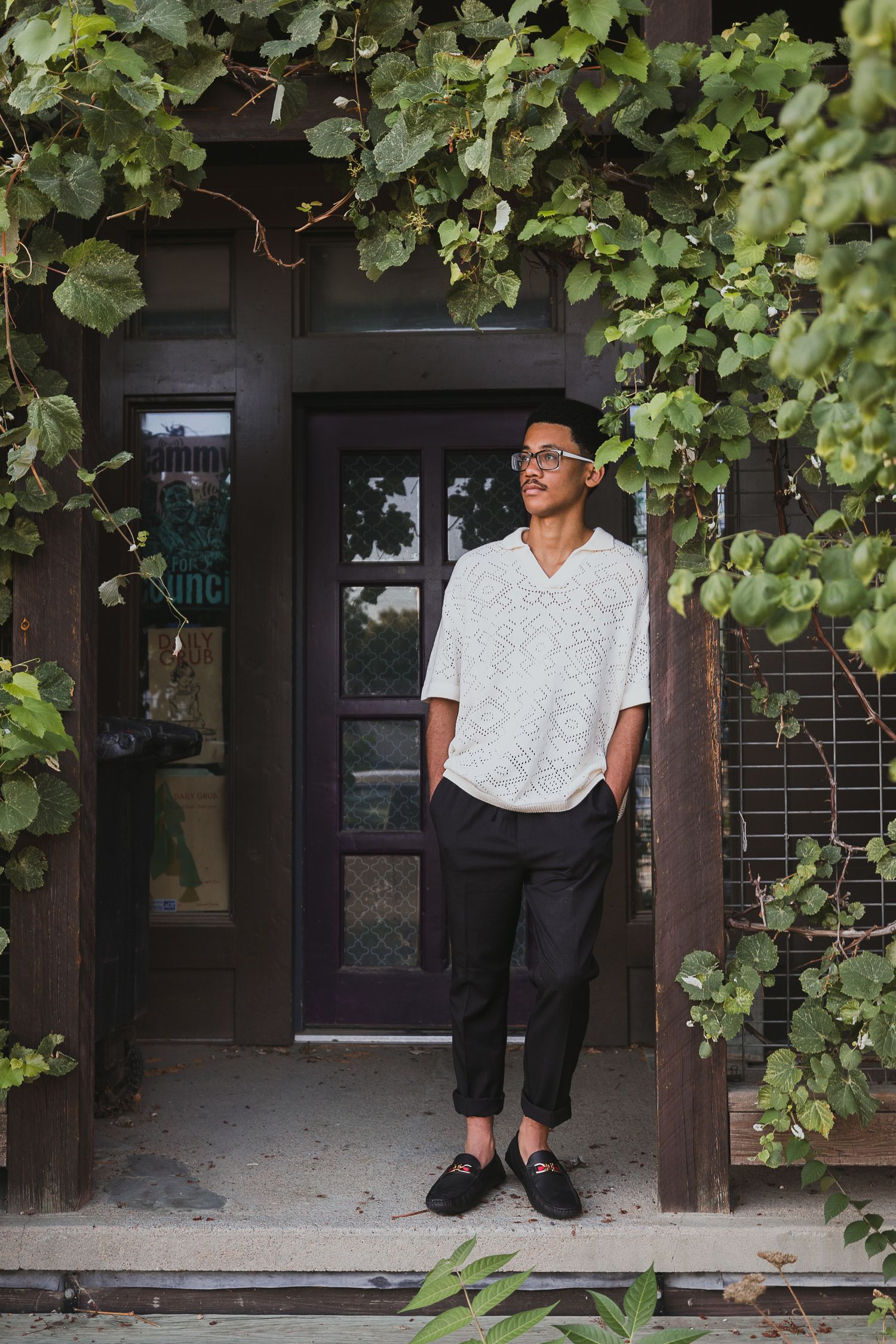 Young man in white shirt and black pants standing by a vine-covered doorway, looking away thoughtfully.