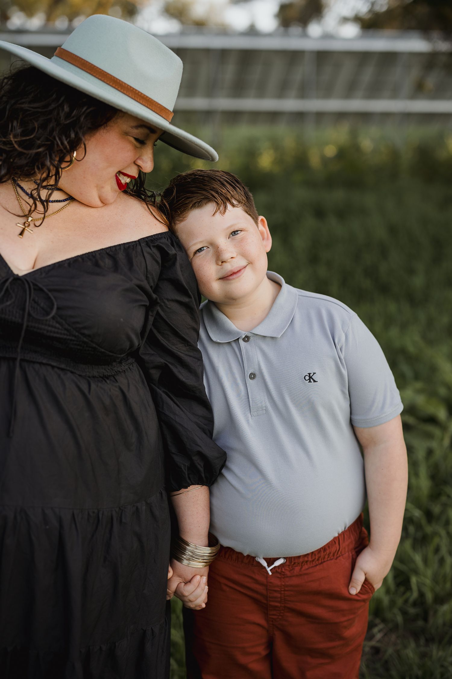 A young boy in a gray polo and red pants holds hands with a woman in a black dress and gray hat outdoors.