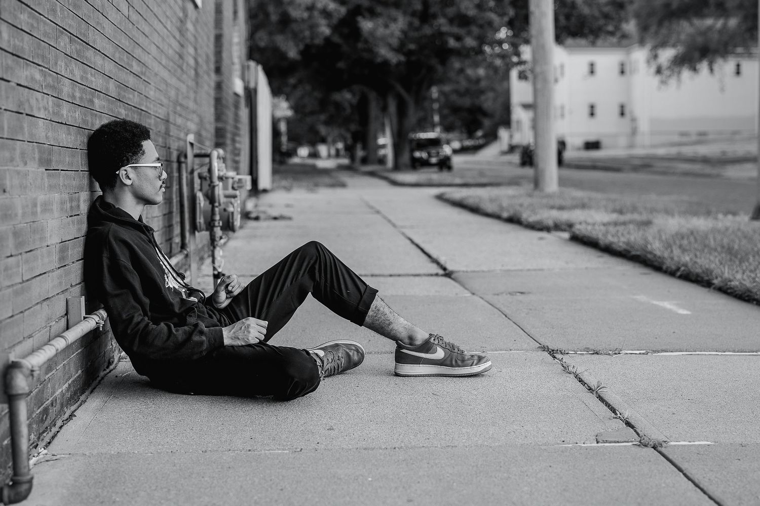 Black and white photo of a young person sitting against a brick wall on a sidewalk, relaxing outdoors.