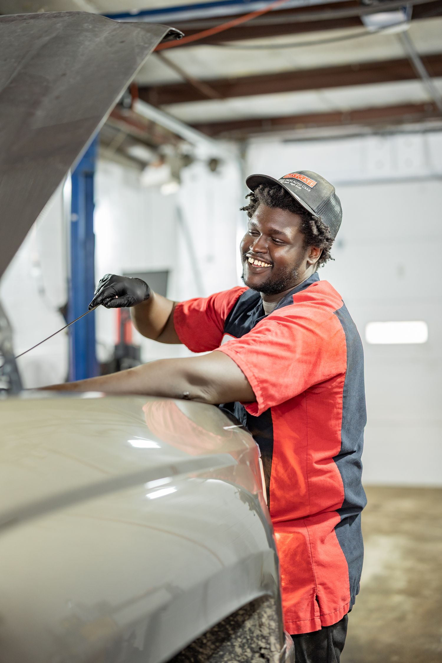 Smiling mechanic in red uniform checking oil dipstick under car hood in auto repair shop.