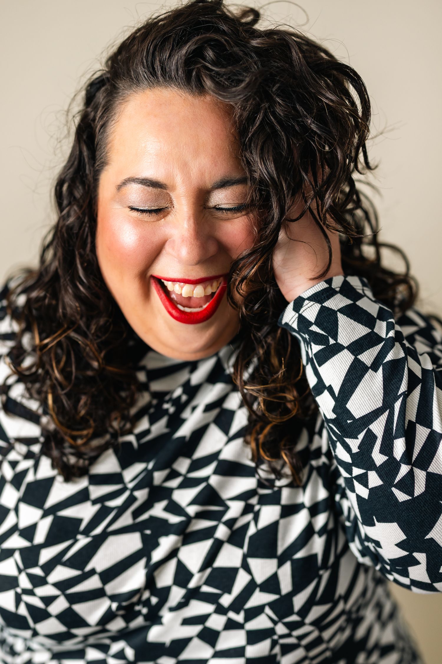 A joyful woman with curly dark hair laughs, wearing a black and white geometric top with bold red lipstick.