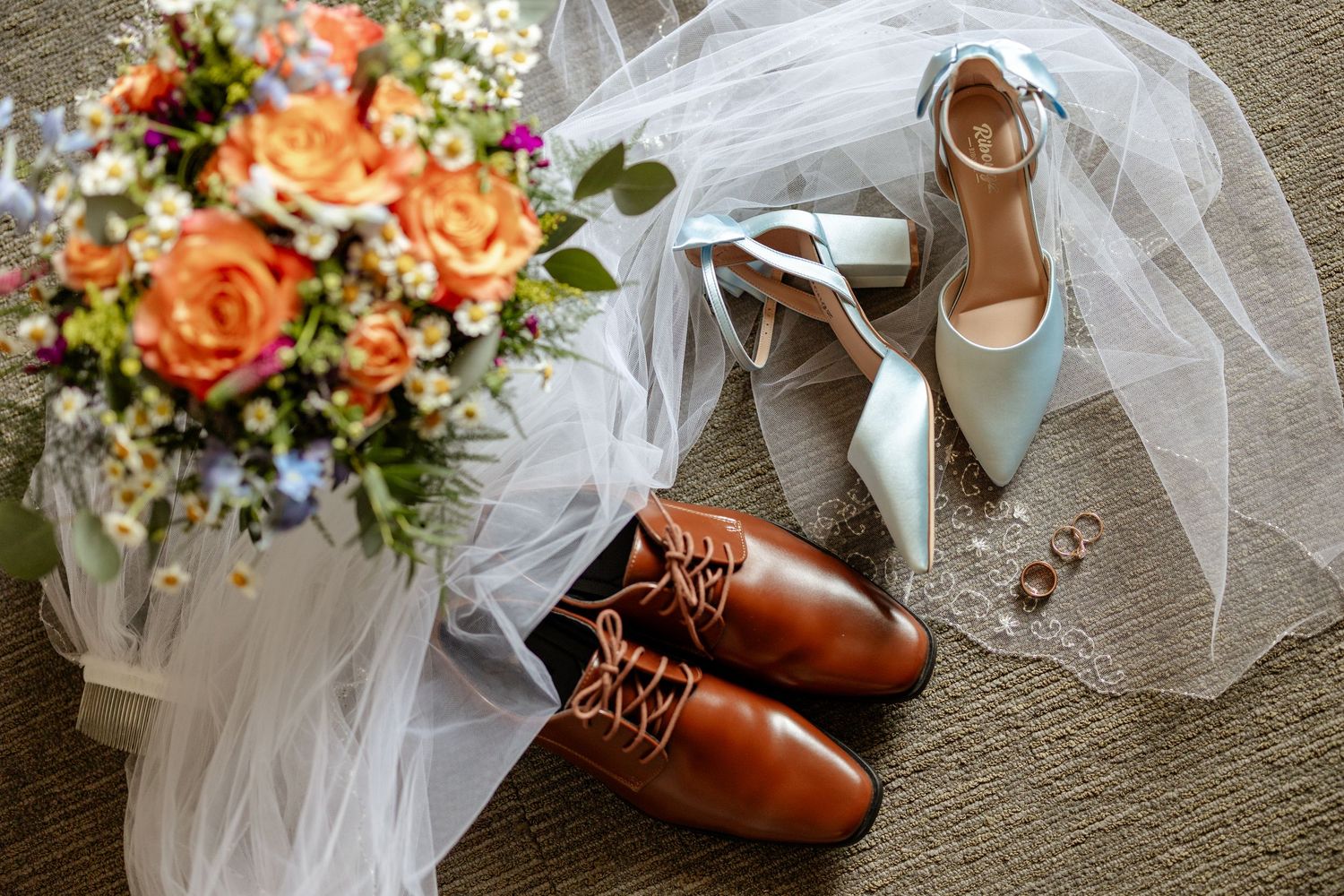 Wedding flat lay featuring orange floral bouquet, brown leather dress shoes, mint heels, gold rings, and white tulle veil.