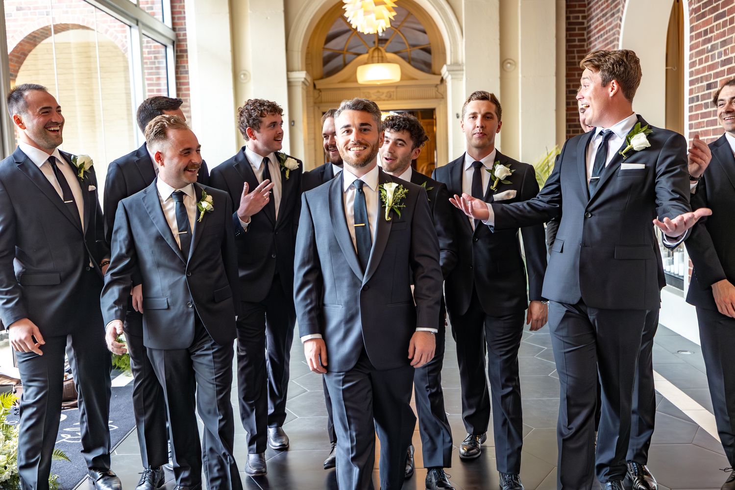 Groom surrounded by smiling groomsmen in dark suits with yellow boutonnieres at an elegant wedding venue.